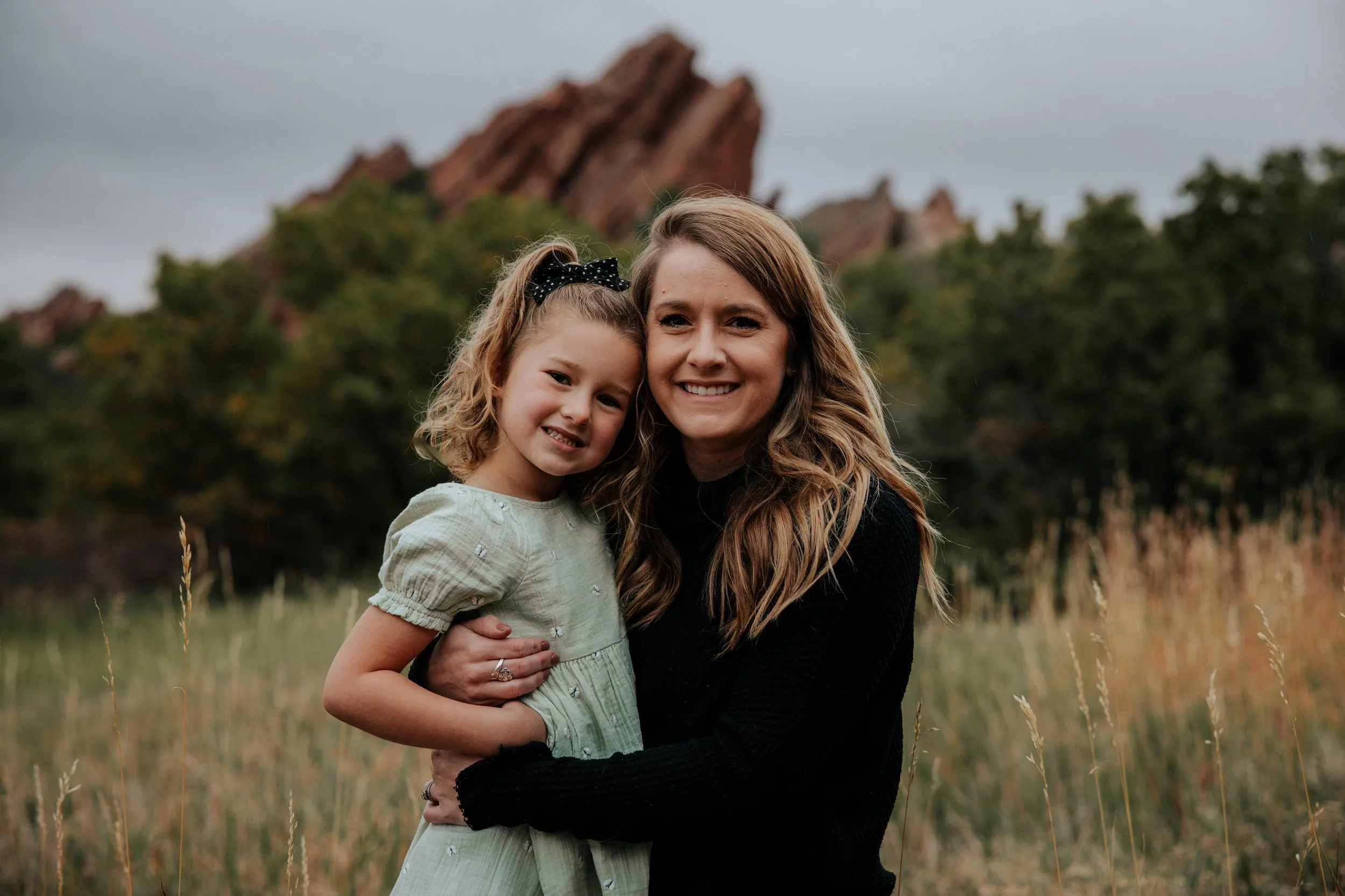 A woman and a young girl smiling and hugging outdoors in a grassy field with trees and rocky formations in the background on a cloudy day.