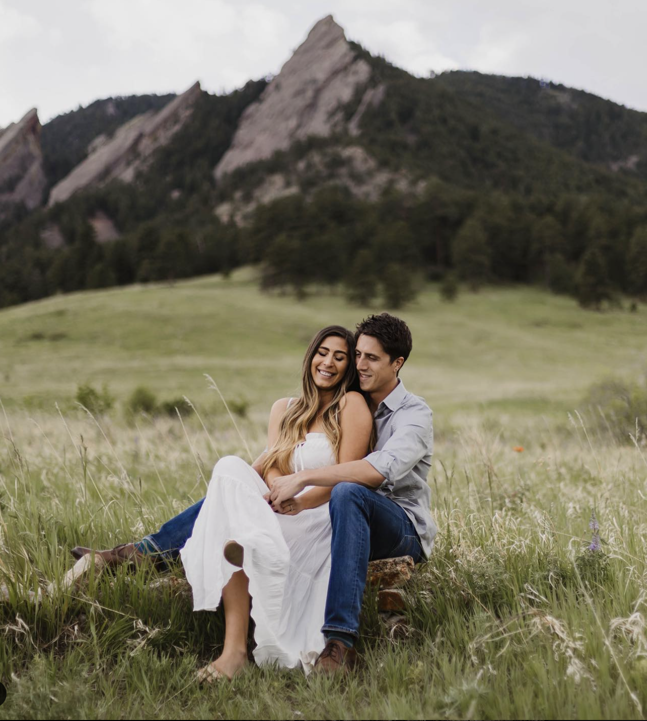 A happy couple sitting on a wood log in a grassy field with mountains in the background, embracing and smiling.