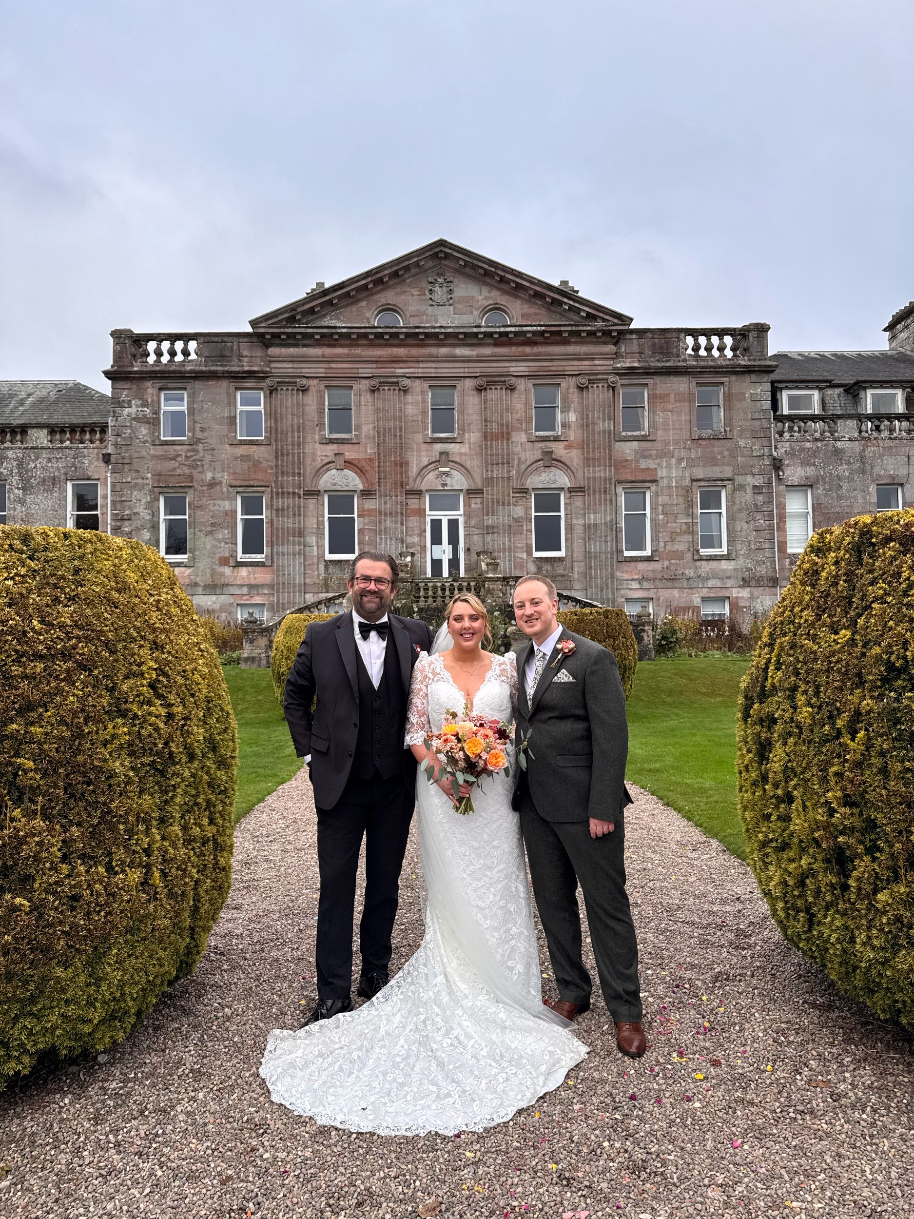 A bride and groom standing outside a historic mansion on their wedding day, with the bride holding a colorful bouquet and dressed in a lace wedding gown, and the groom in a dark suit. The mansion has a stone facade, multiple windows, and decorative e