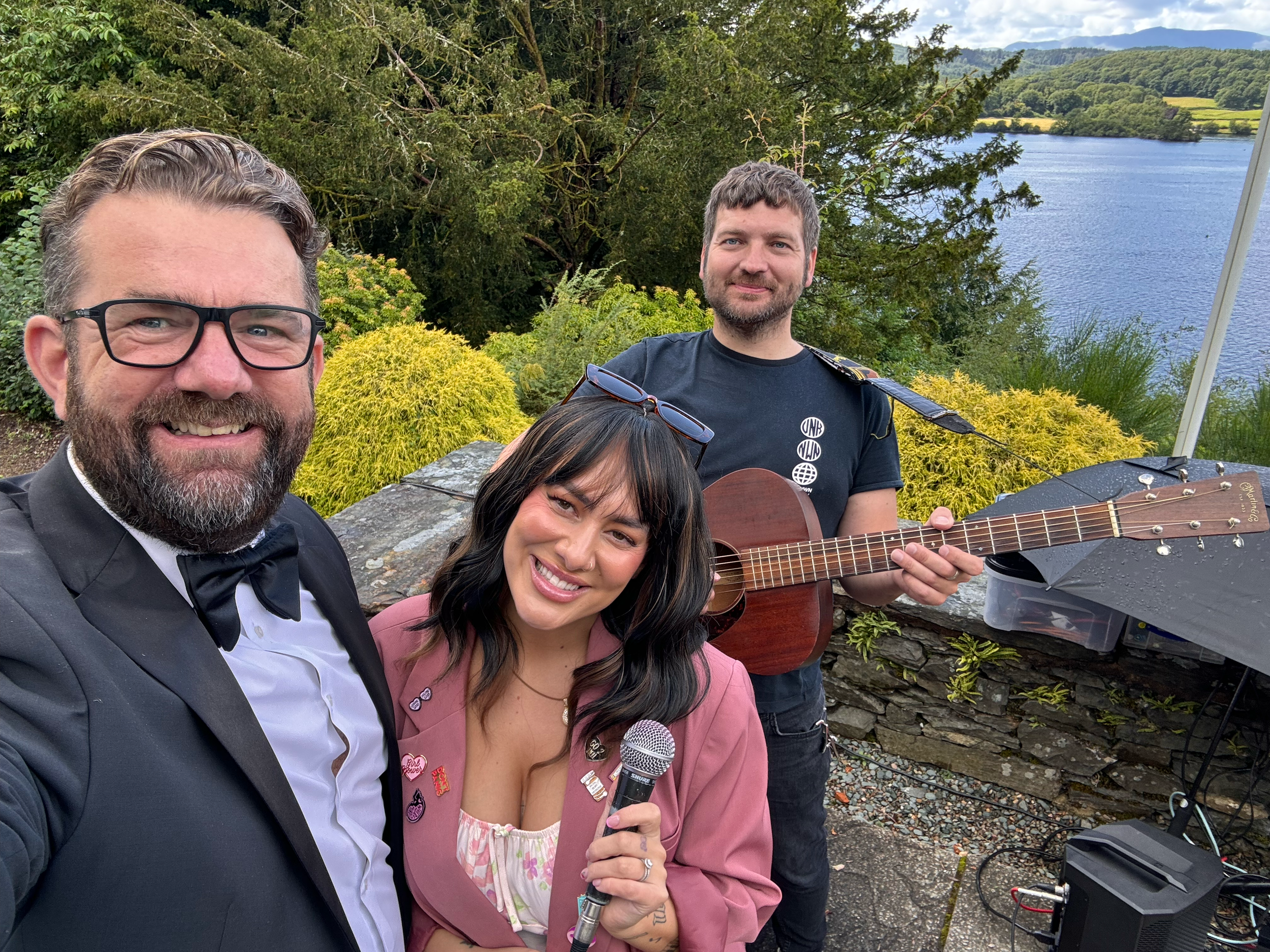 Three people posing outdoors; a woman in a pink jacket holding a microphone and a guitar, smiling, with a man with glasses and a beard beside her, and another man on the left side of the image with gray hair and a beard. The group is surrounded by gr