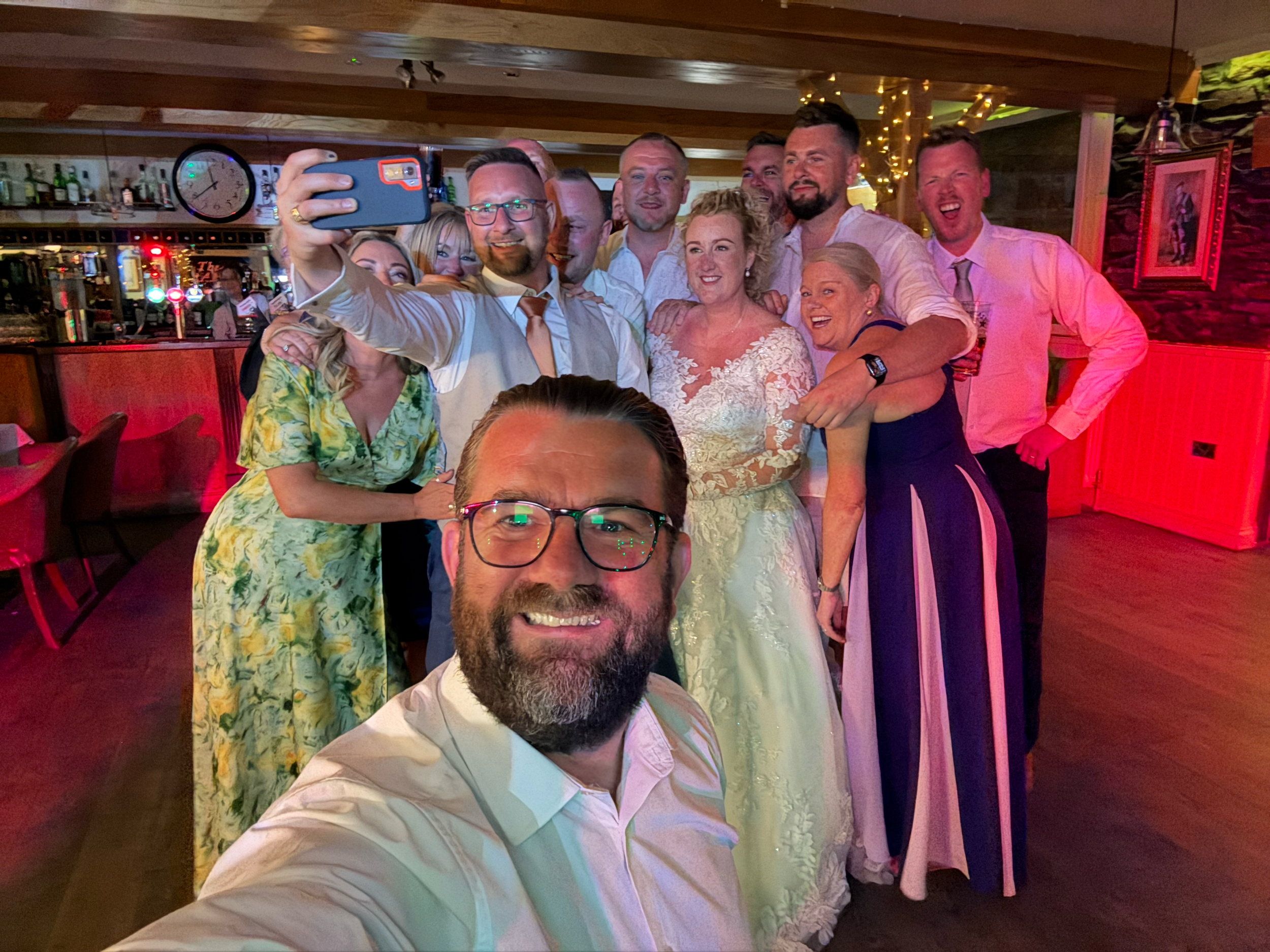 Group of people taking a selfie in a cozy indoor setting with warm lighting and a bar in the background, all smiling and posing happily.