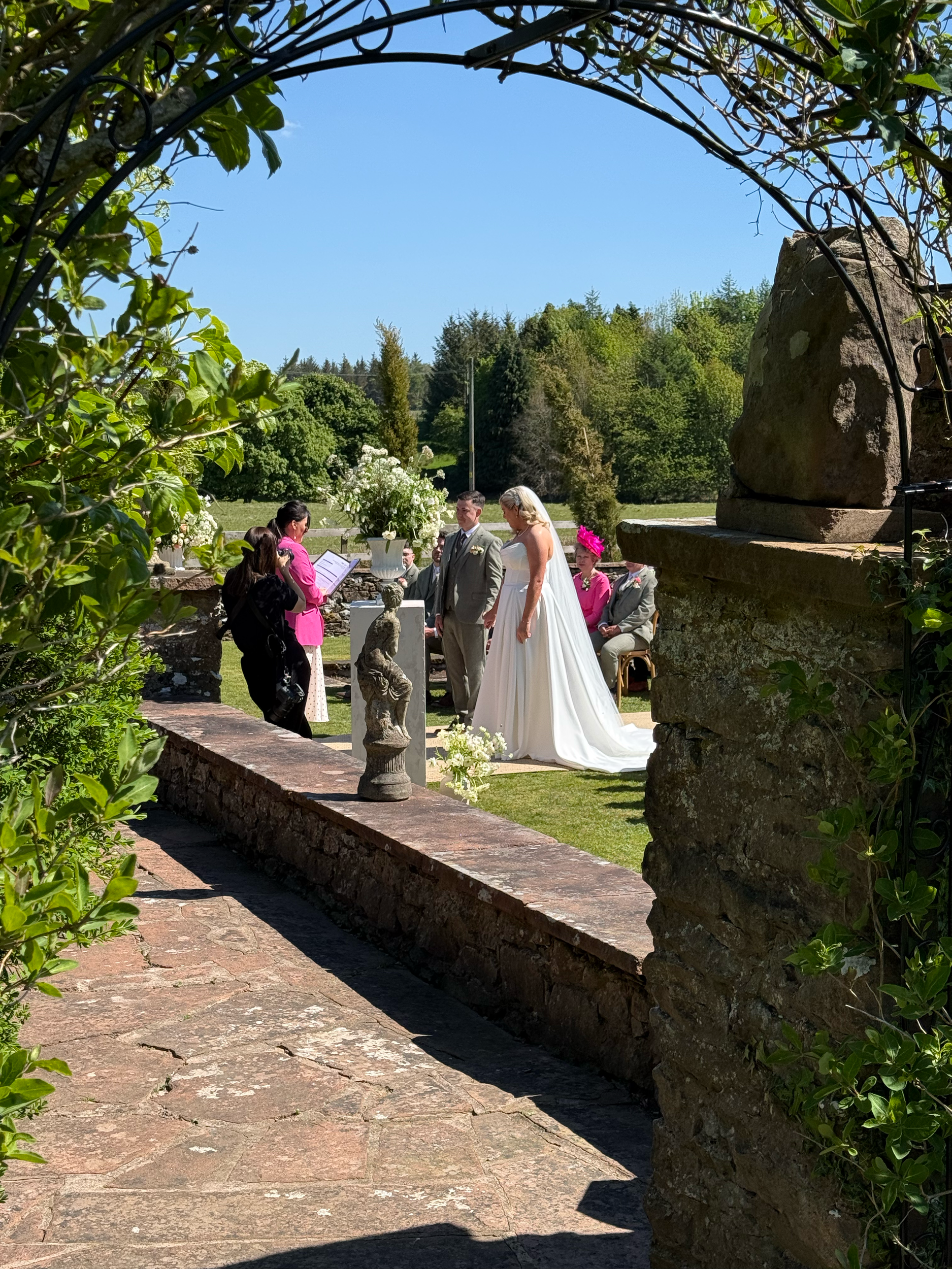 A wedding ceremony outdoors with a bride in a white dress and a groom in a light-colored suit standing behind a table adorned with flowers, under a stone arch. Several guests are seated and standing nearby. The setting is lush with greenery and trees