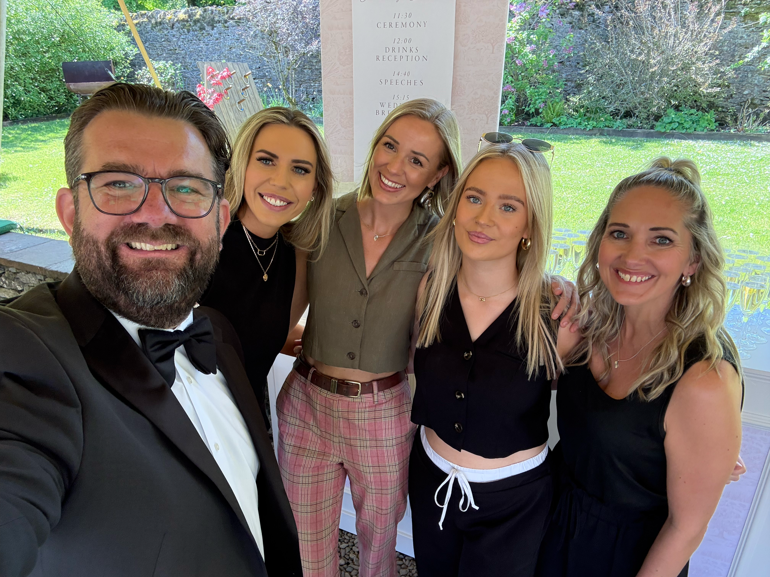 Group of six people, five women and one man, dressed in formal attire, smiling for a photo outdoors with greenery in the background.