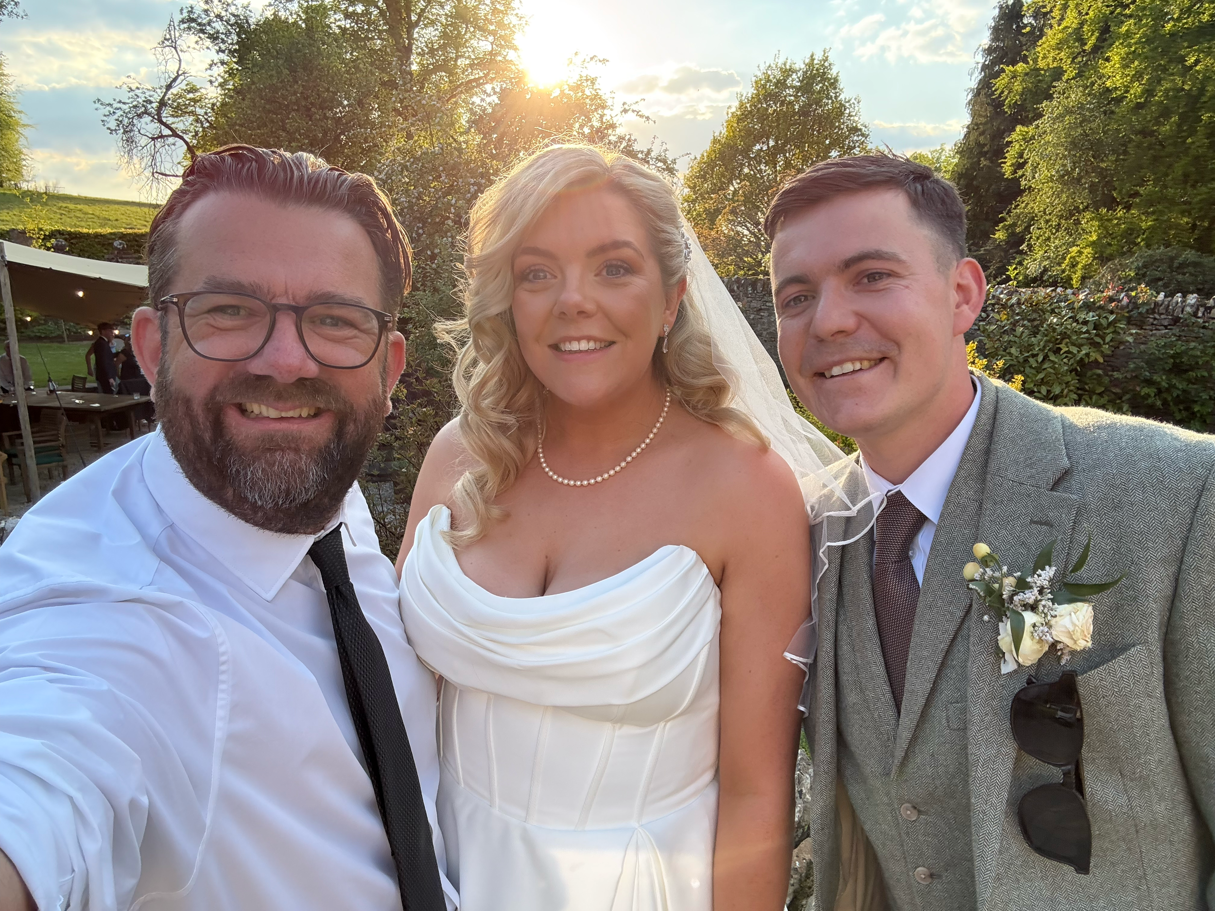 A newlywed couple and a man pose for a photo outdoors during sunset. The bride wears a white wedding dress and pearl necklace, the groom wears a grey suit with sunglasses, and the man on the right wears glasses and a white shirt.