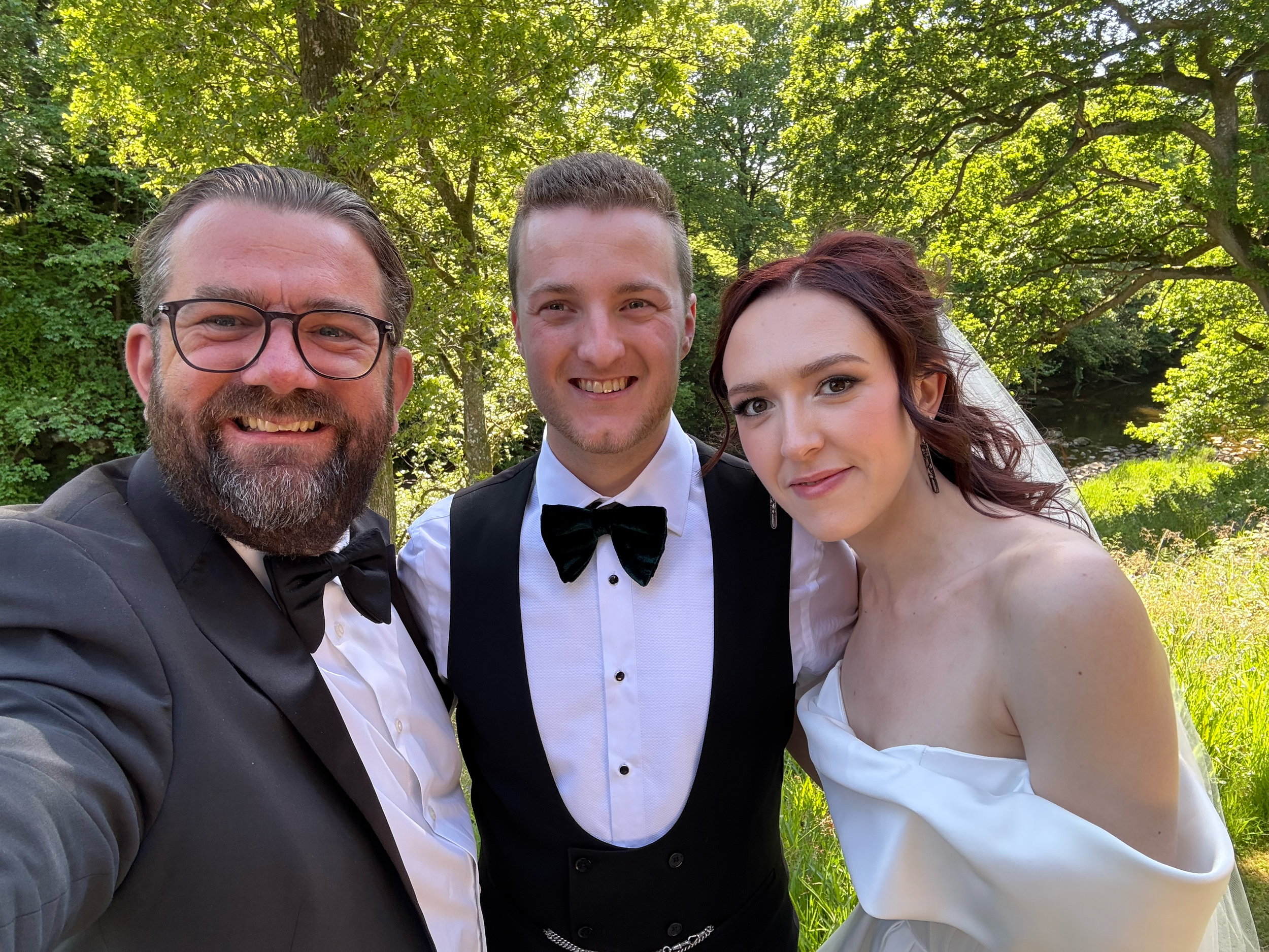 Three people dressed in wedding attire smiling and standing outdoors under green trees.