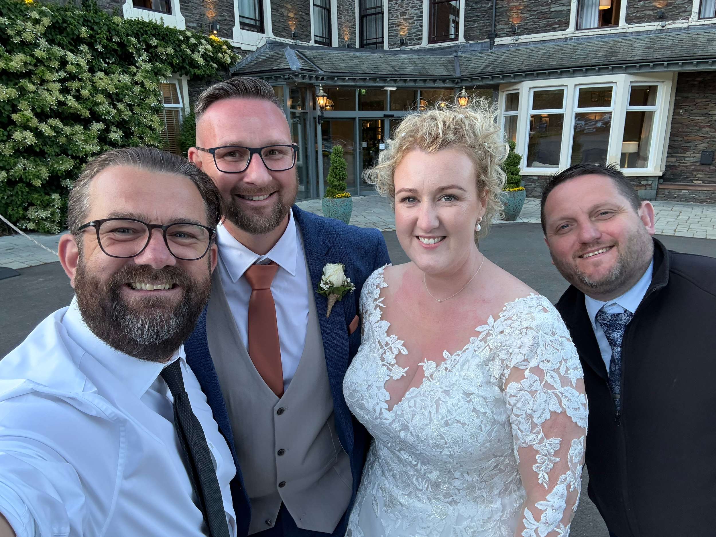 Group of five people, including a bride in a white lace wedding dress and groom in a blue suit, taking a selfie outside a building with brick and glass windows, with a large bush of white flowers in the background.