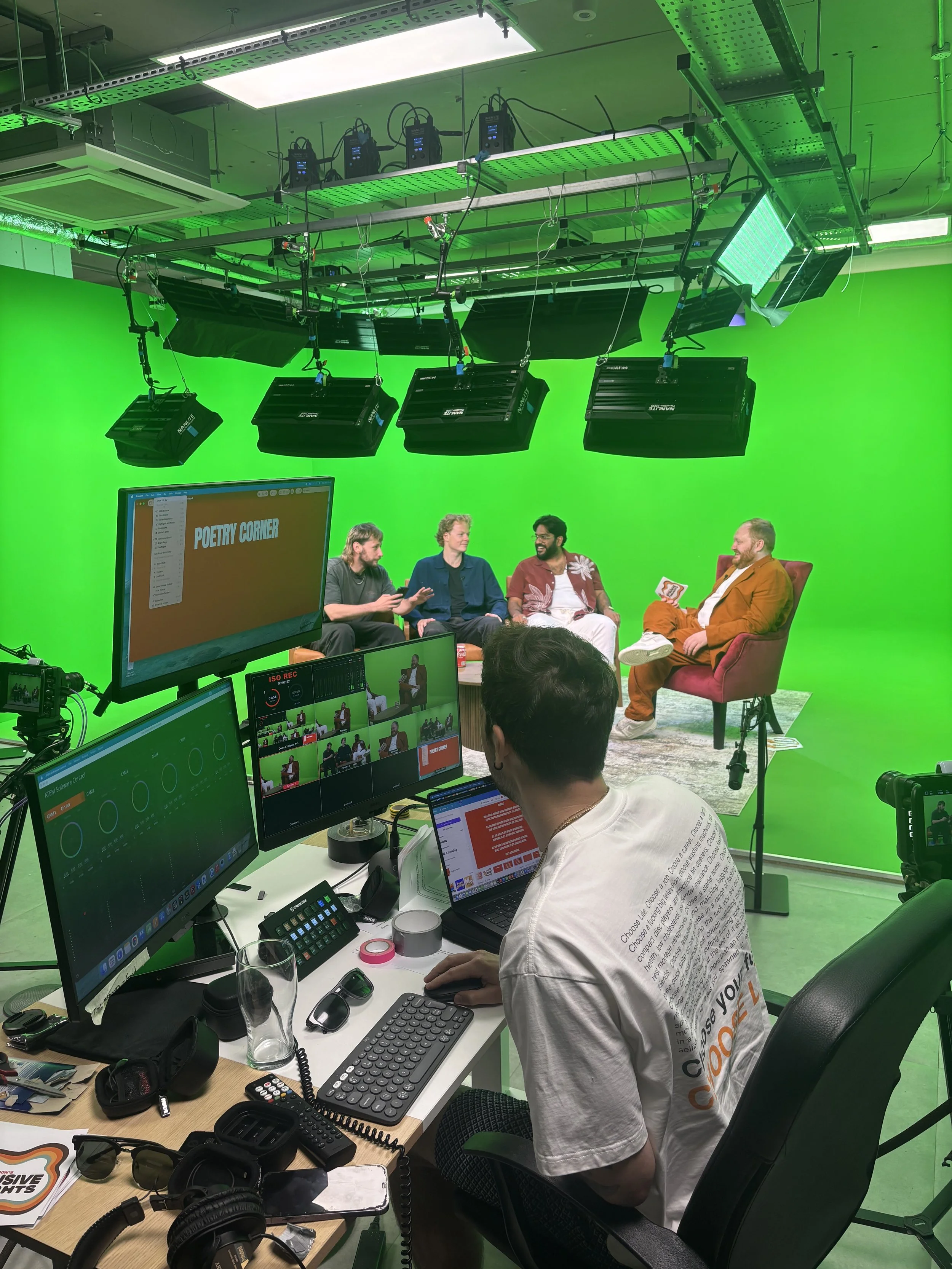 A television studio with a green screen, of people sitting on chairs, engaged in conversation. There are multiple lighting and camera equipment, screens, and a person working at a desk with monitors, keyboards, and various electronic devices.
