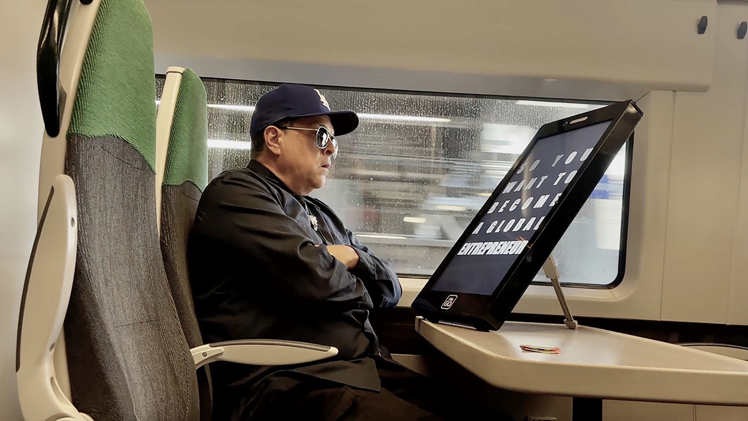 A man wearing sunglasses, a navy cap, and a black jacket is sitting on a train, with arms crossed, looking to the right. He is next to a window, and a large digital screen displays text behind him.