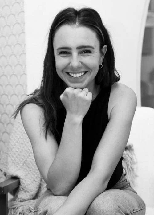 Carly Pollack smiling, with long dark hair, wearing a sleeveless top, sitting with one arm resting on her knee and her fist near her chin, in an indoor setting.
