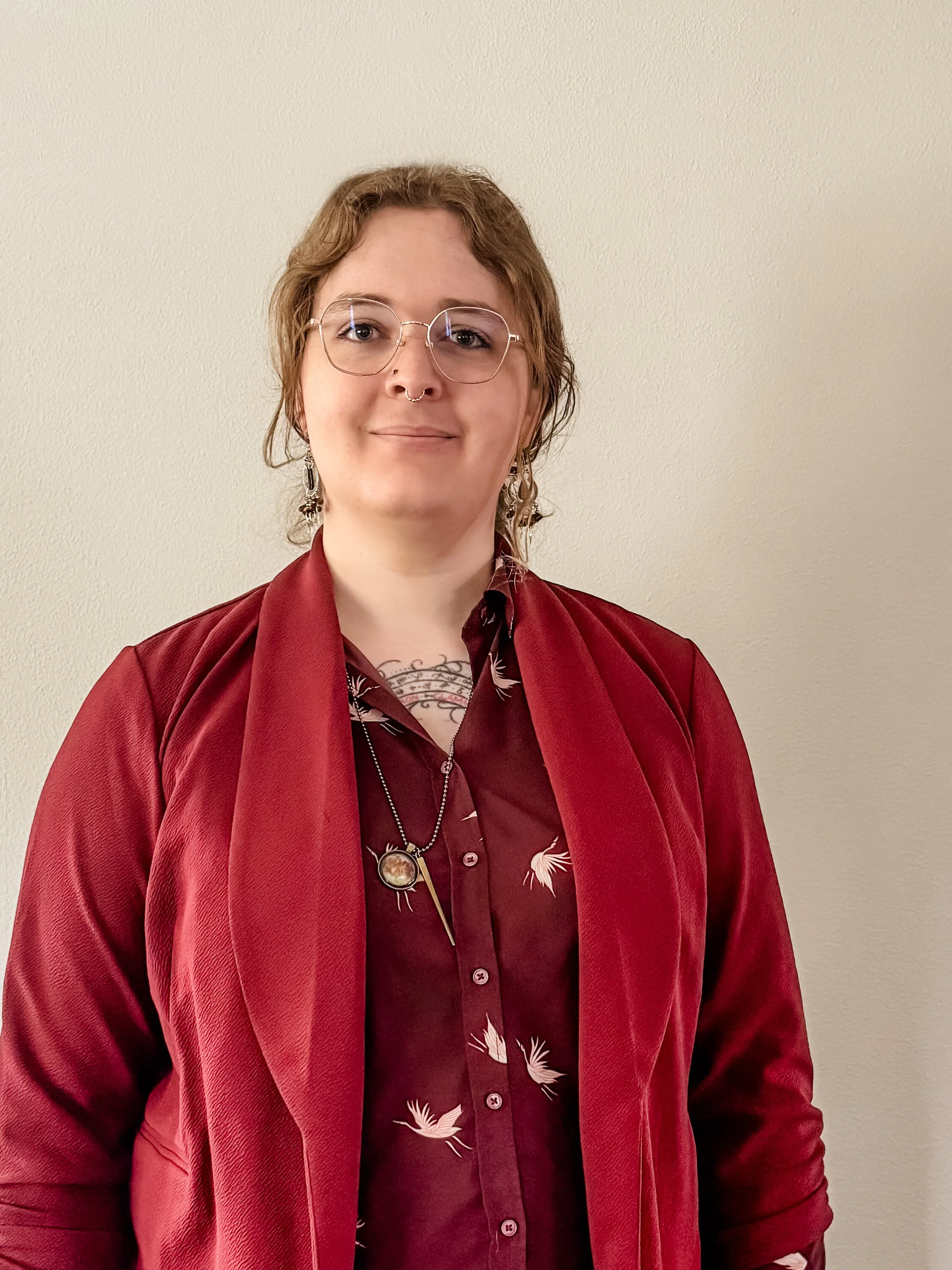 A woman with light skin, curly red hair, glasses, and a septum piercing, wearing a maroon shirt with a white bird pattern and a matching maroon jacket, standing against a plain light beige wall.