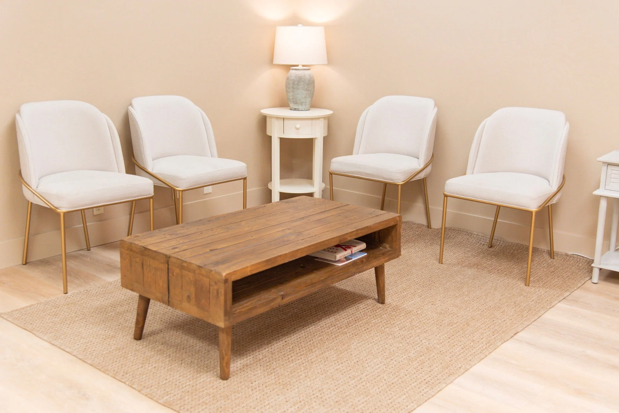 Waiting room with four white chairs with gold legs arranged in a semi-circle around a round coffee table. The table has a small potted plant, magazines, and a clear plastic stand. There is a beige rug underneath the table and light wooden flooring. A small white side table with a lamp and a small plant is against the wall, creating a cozy and modern waiting area.