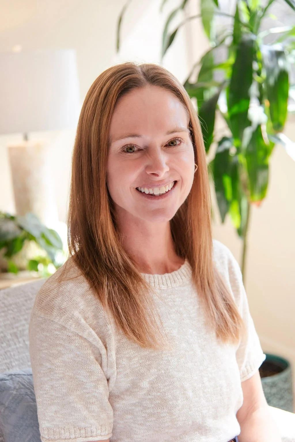 A woman with reddish-brown hair smiling indoors, with houseplants and a lamp in the background.