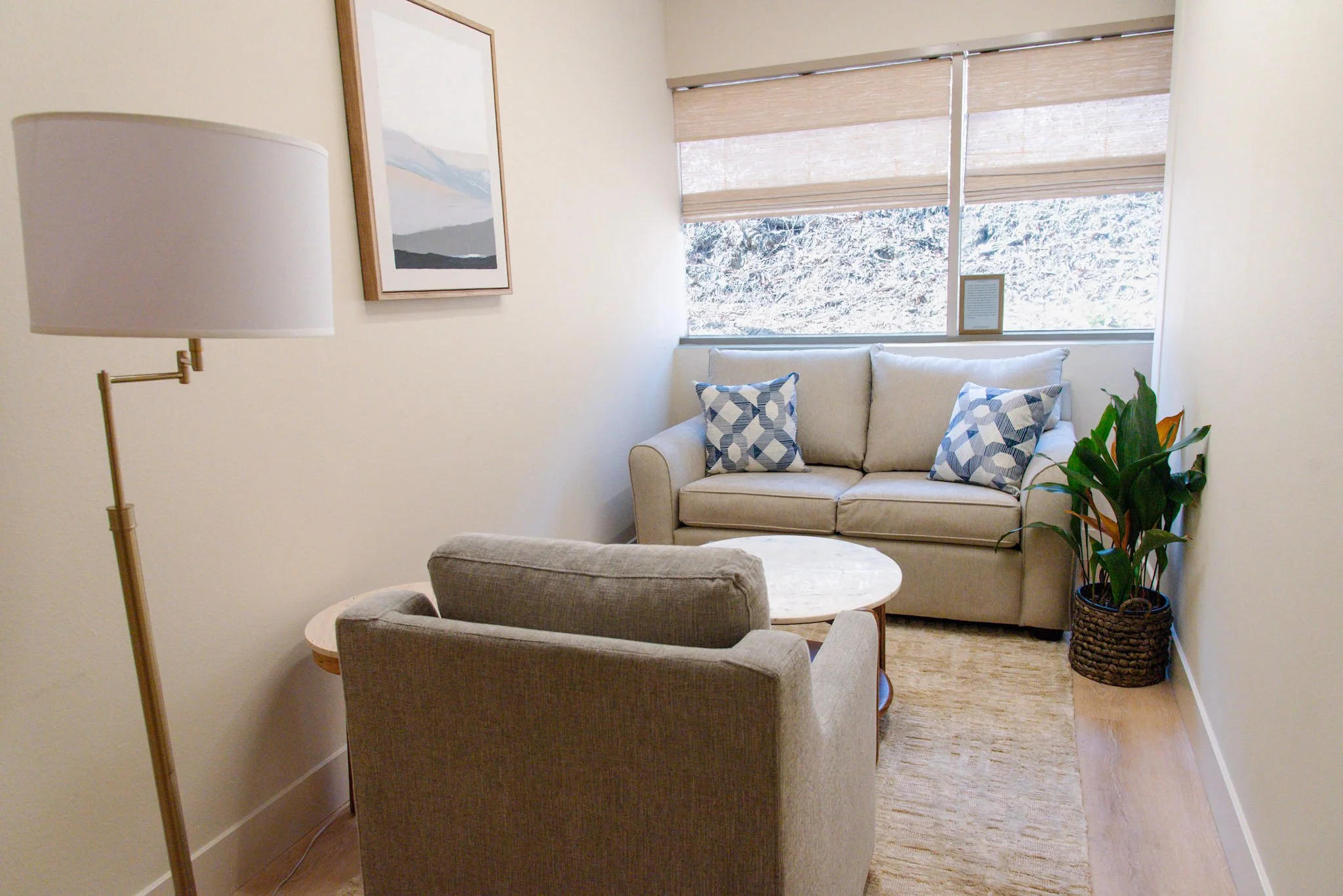 Living room with a beige armchair, a gray couch with two patterned pillows, a round wooden coffee table, a potted plant, a framed landscape art piece, and a window with beige blinds overlooking greenery.