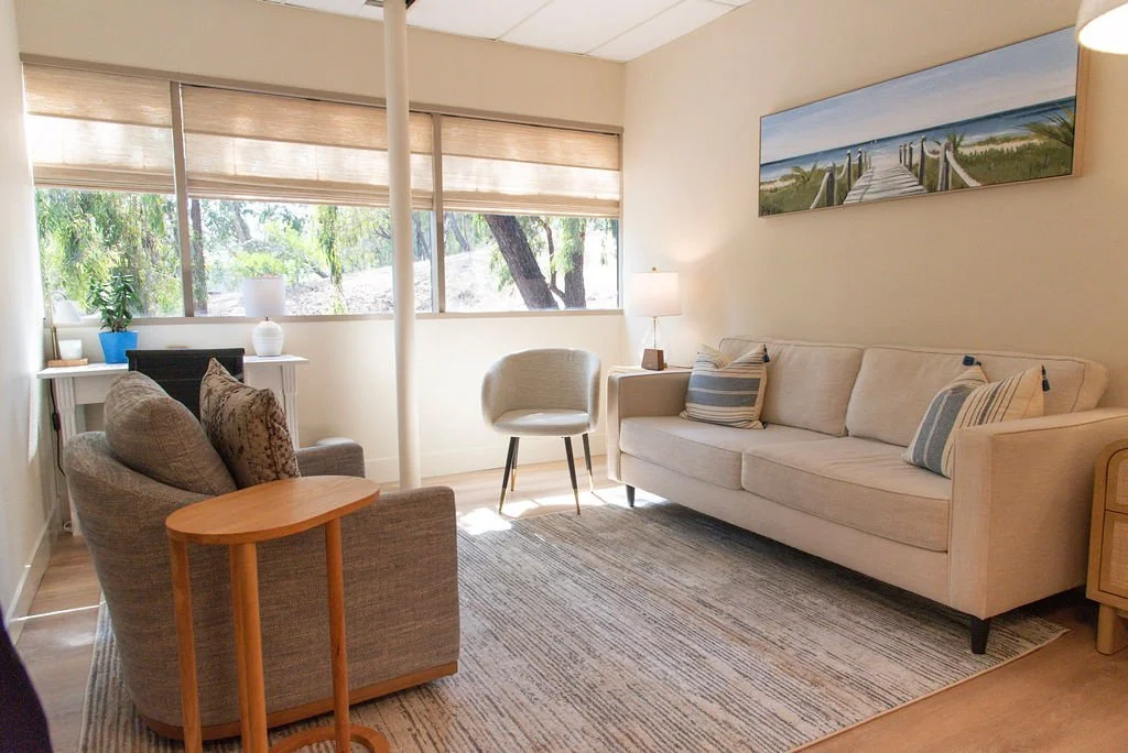 A cozy living room with beige walls and wooden flooring, featuring a white sofa with striped pillows, an armchair, a small wooden side table, a white side table with a potted plant, large window with blinds showing greenery outside, a floor lamp, a landscape painting of a pier on the wall, and a cream area rug.