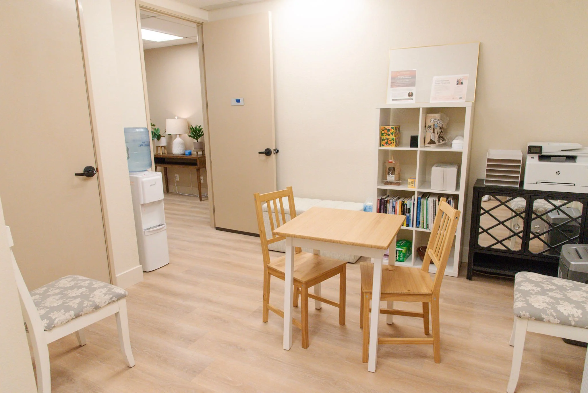 A small dining area with a white oval table and four white chairs with floral cushions. In the background, there is a microwave, a small refrigerator, and a water dispenser with bottled water. The floor is light wood, and the walls are beige.