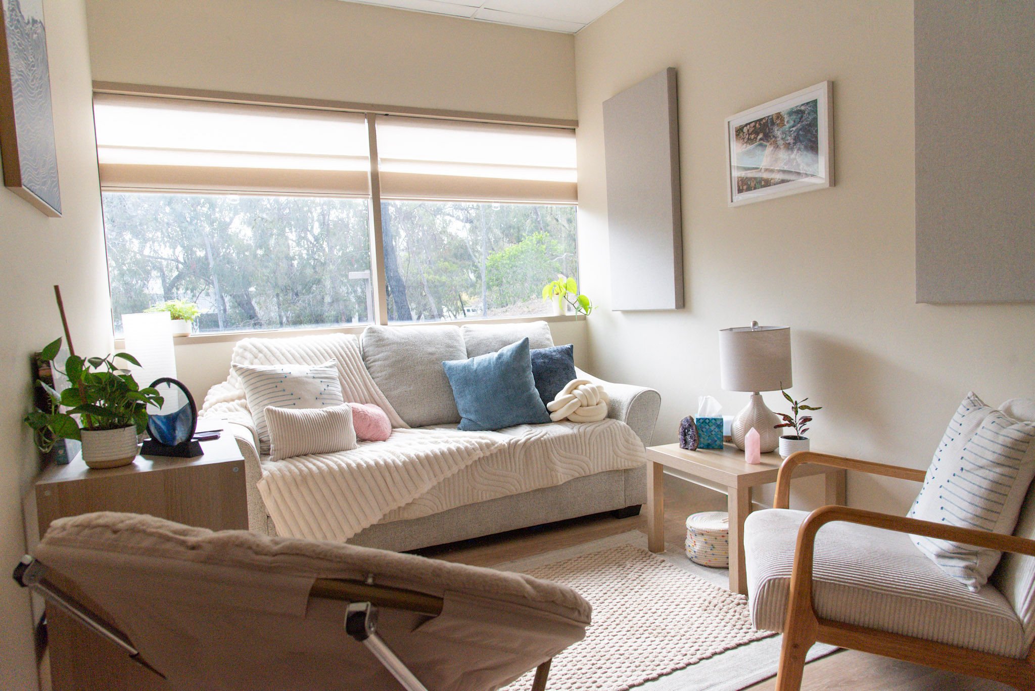 Living room with a gray sofa, pillows, a beige armchair, wooden side table, carpet, window view of trees and sky, and indoor plants.