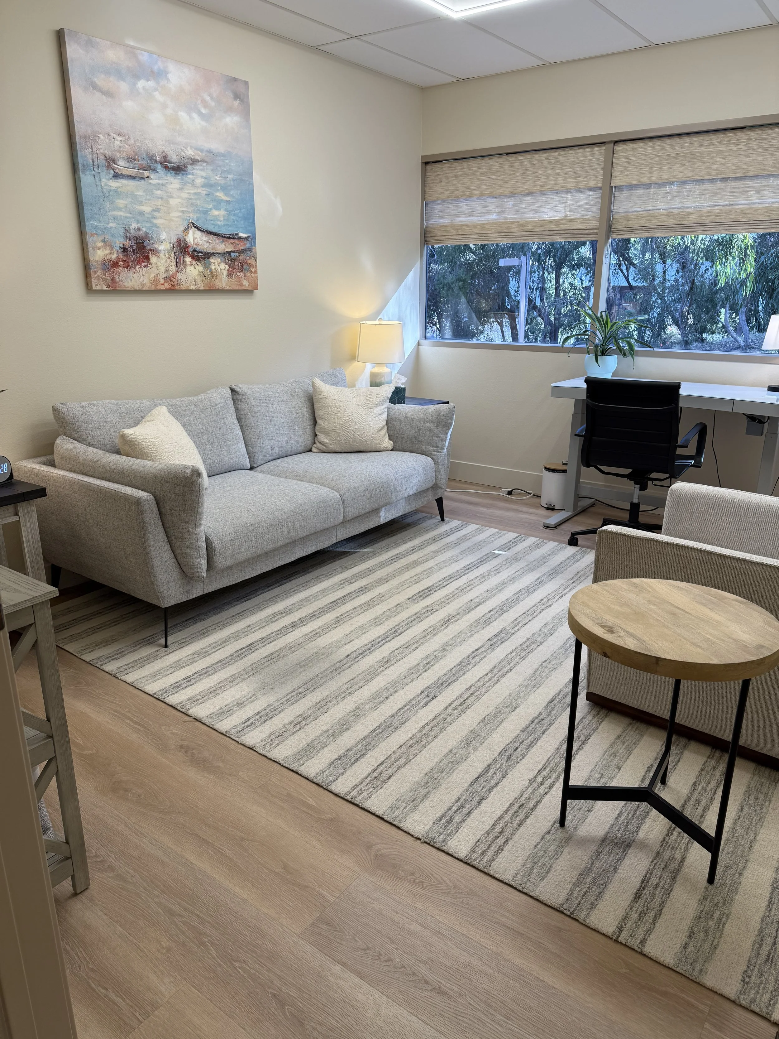 A cozy living room with a light gray sofa, striped rug, round wooden side table, and a workspace near a large window with blinds and greenery outside.
