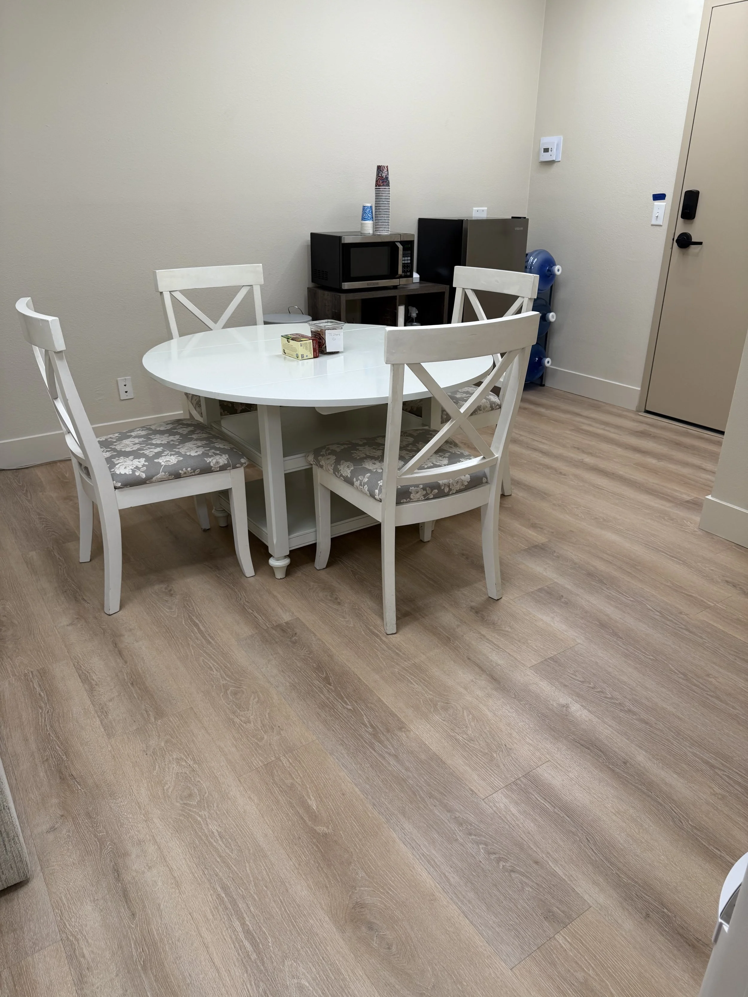 A small dining area with a white oval table and four white chairs with floral cushions. In the background, there is a microwave, a small refrigerator, and a water dispenser with bottled water. The floor is light wood, and the walls are beige.