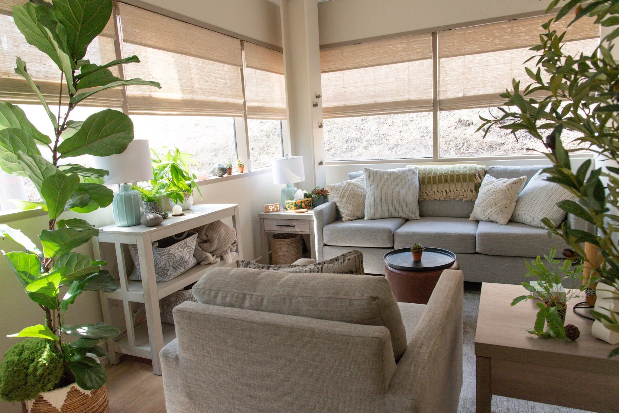 A cozy therapy room with a beige sofa, a light-colored armchair, a small rug, a large window with blinds, potted plants, and decorative pillows.