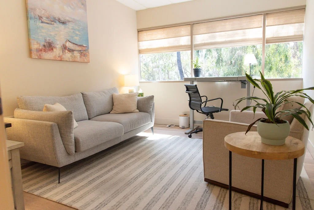 A cozy therapy room with a light gray sofa, striped rug, round wooden side table, and a workspace near a large window with blinds and greenery outside.