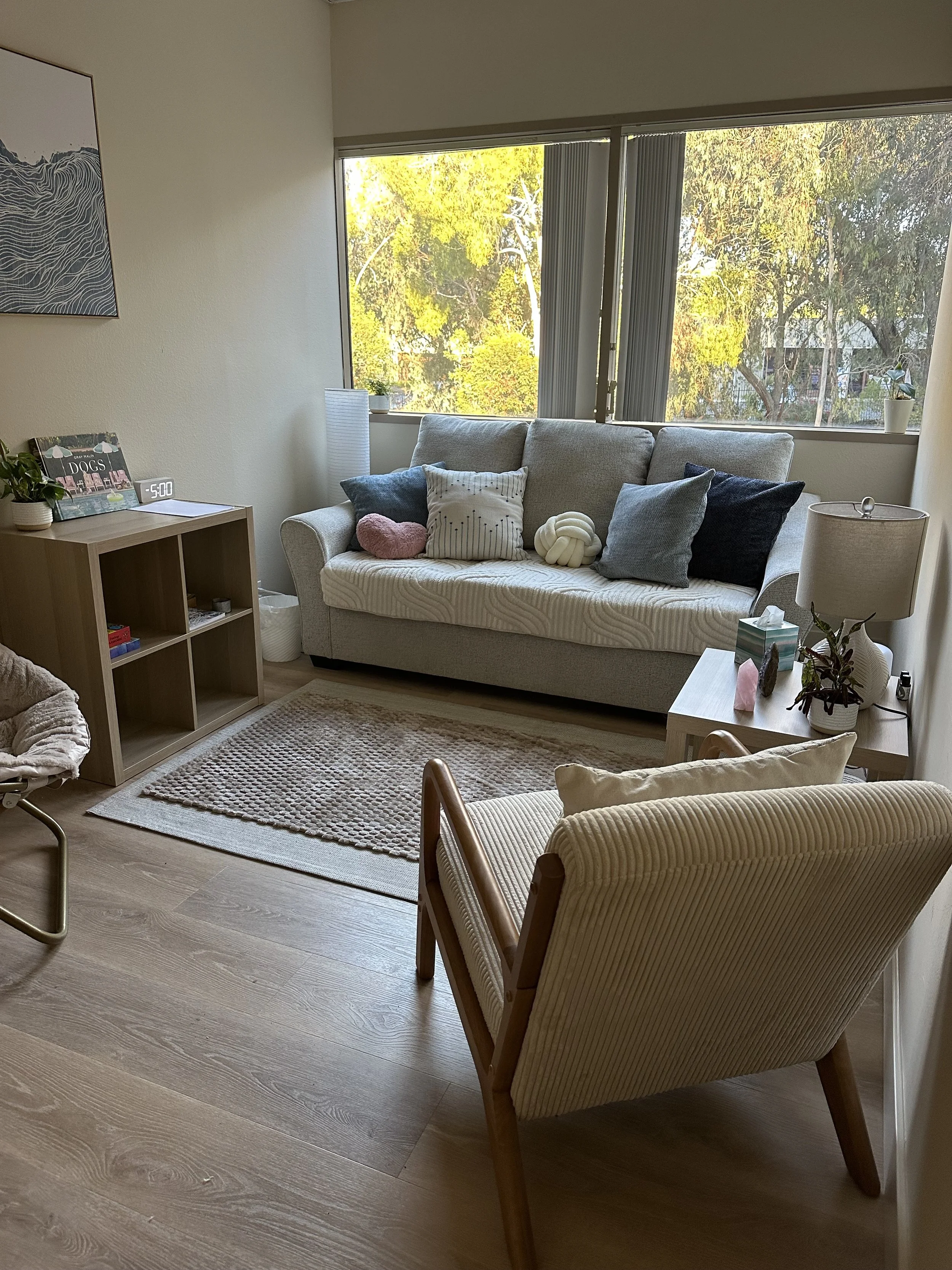 Living room with a gray sofa, pillows, a beige armchair, wooden side table, carpet, window view of trees and sky, and indoor plants.