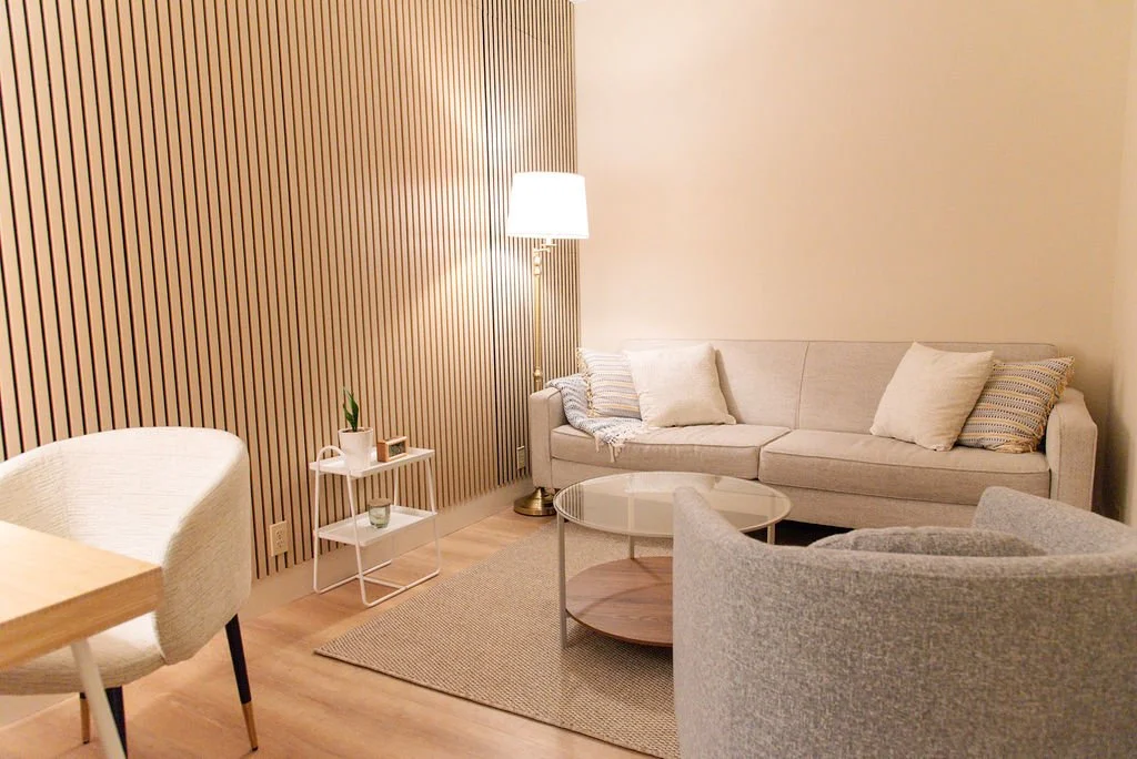 Small living room with light gray couch, two patterned pillows, a wooden coffee table, a gray armchair, two white side tables, a potted plant, and a seaside photograph on the wall.