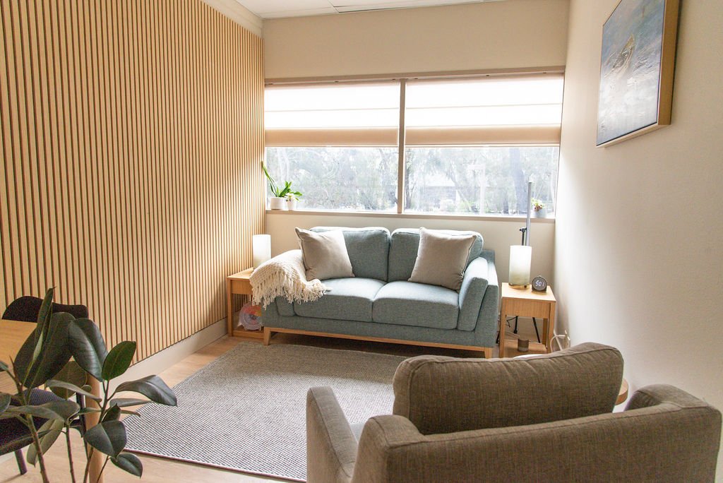 A cozy living room with a teal sofa and two beige armchairs arranged around a small wooden side table and a round wooden coffee table. Large windows with beige blinds let in natural light, and there is a painting on the wall.