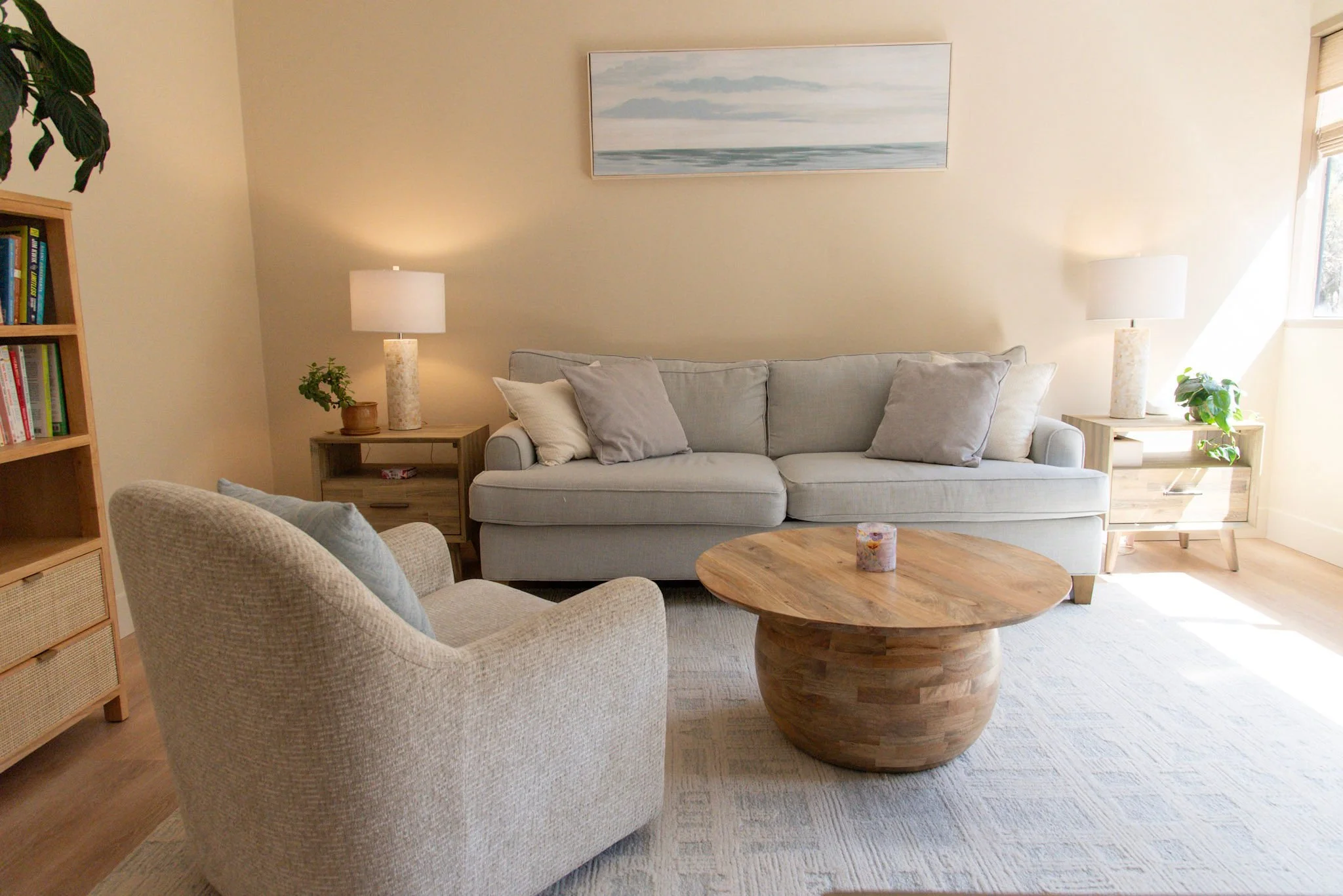 Living room with a light gray sofa, four throw pillows, a side table with a lamp, a beige armchair with a patterned throw blanket, a round coffee table, large windows with beige blinds, a potted plant, and a framed picture of a seascape on the wall.