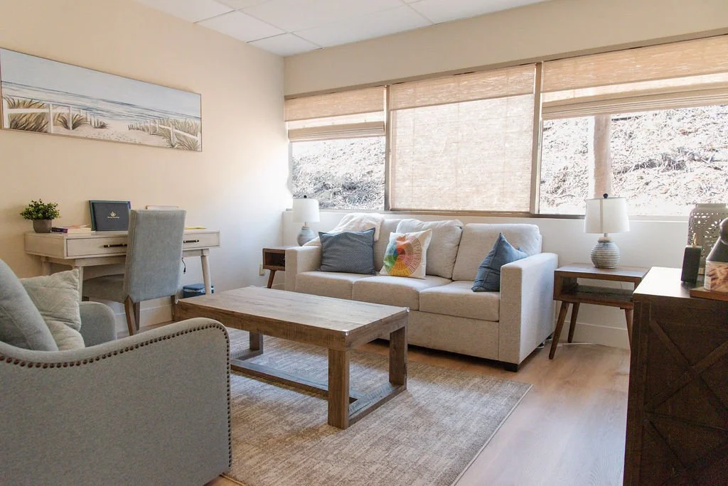 A therapy room with a beige sofa, blue and beige pillows, a wooden coffee table, two side tables with lamps, a gray armchair, a desk with a chair, and a window with shades showing greenery outside.