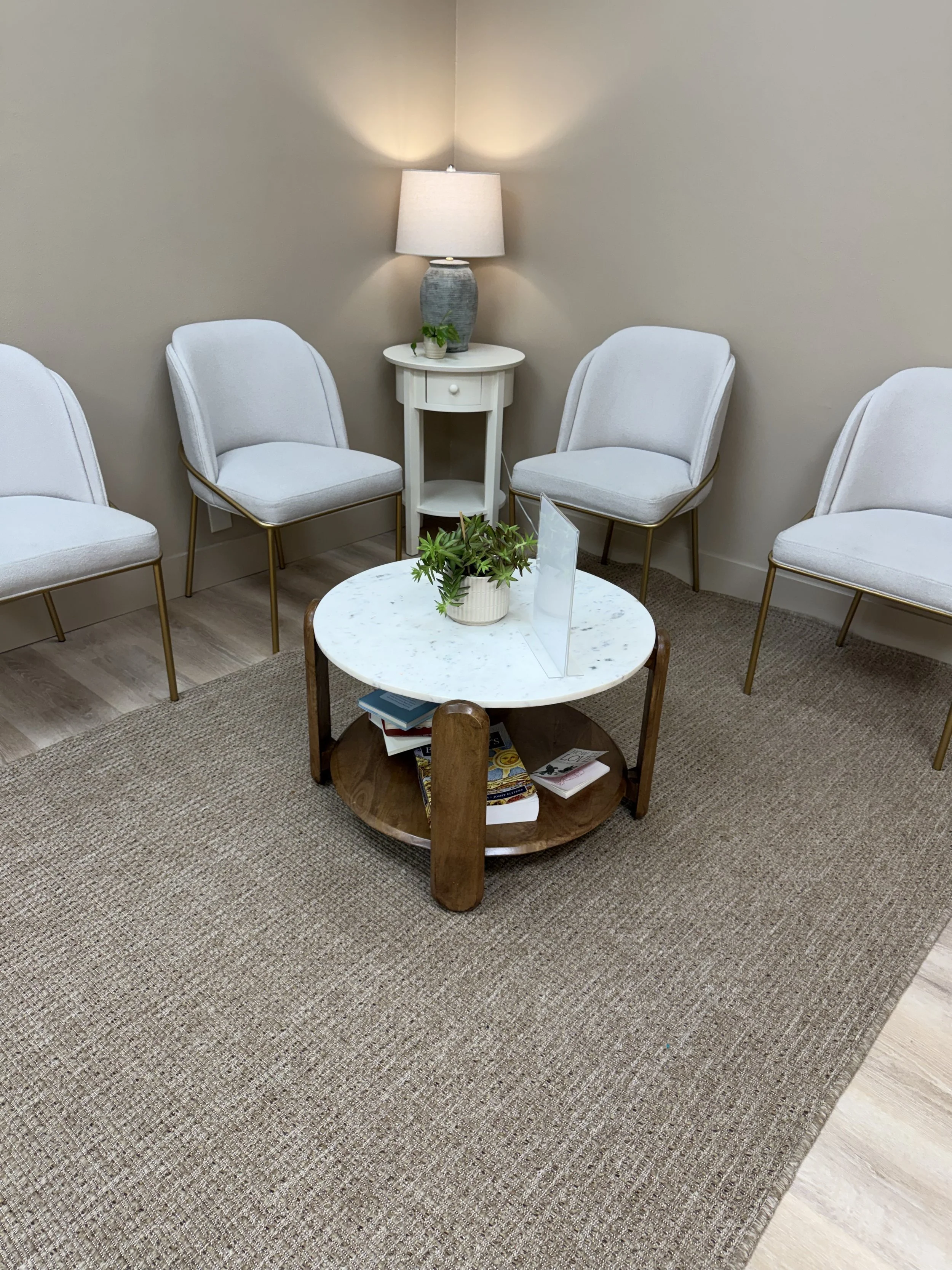 Waiting room with four white chairs with gold legs arranged in a semi-circle around a round coffee table. The table has a small potted plant, magazines, and a clear plastic stand. There is a beige rug underneath the table and light wooden flooring. A small white side table with a lamp and a small plant is against the wall, creating a cozy and modern waiting area.