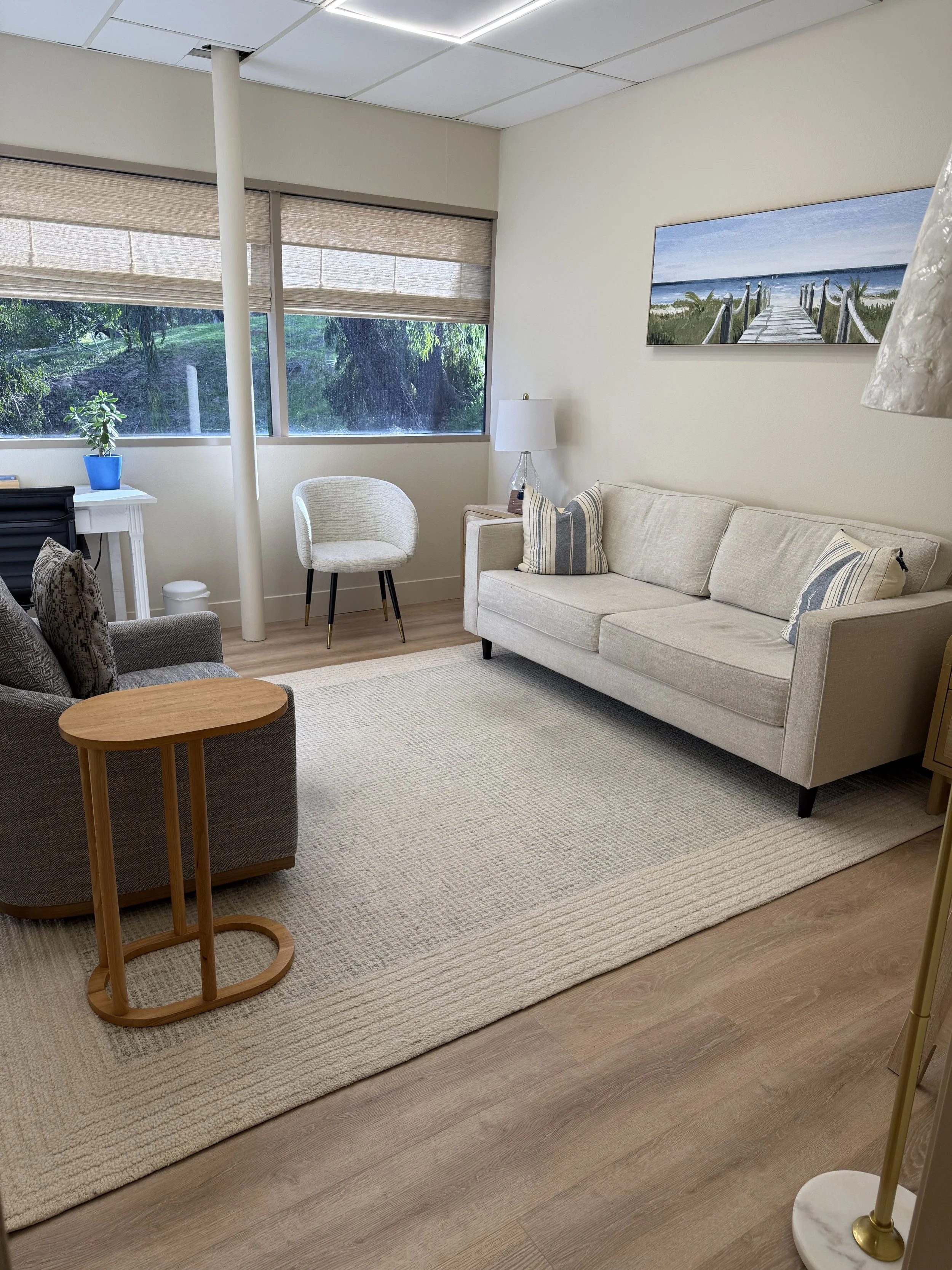 A cozy living room with beige walls and wooden flooring, featuring a white sofa with striped pillows, an armchair, a small wooden side table, a white side table with a potted plant, large window with blinds showing greenery outside, a floor lamp, a landscape painting of a pier on the wall, and a cream area rug.