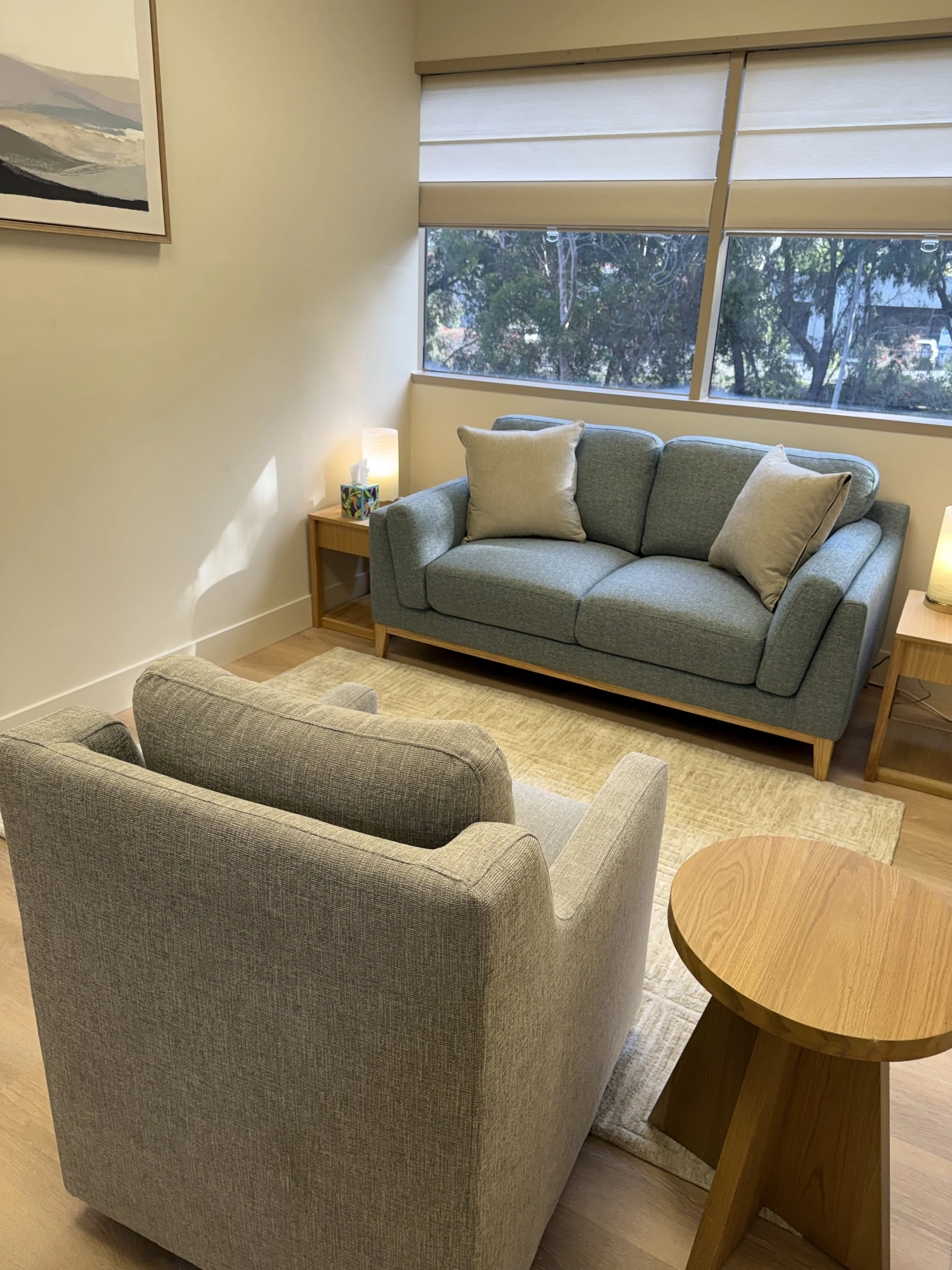 A cozy living room with a teal sofa and two beige armchairs arranged around a small wooden side table and a round wooden coffee table. Large windows with beige blinds let in natural light, and there is a painting on the wall.