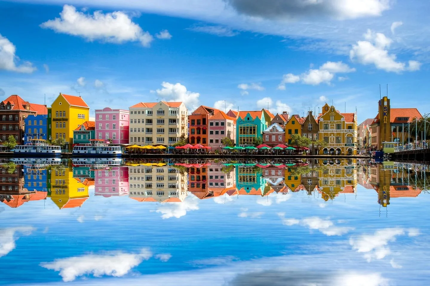 Colorful buildings lining a waterfront with boats and umbrellas, reflected in the water and under a blue sky with clouds.
