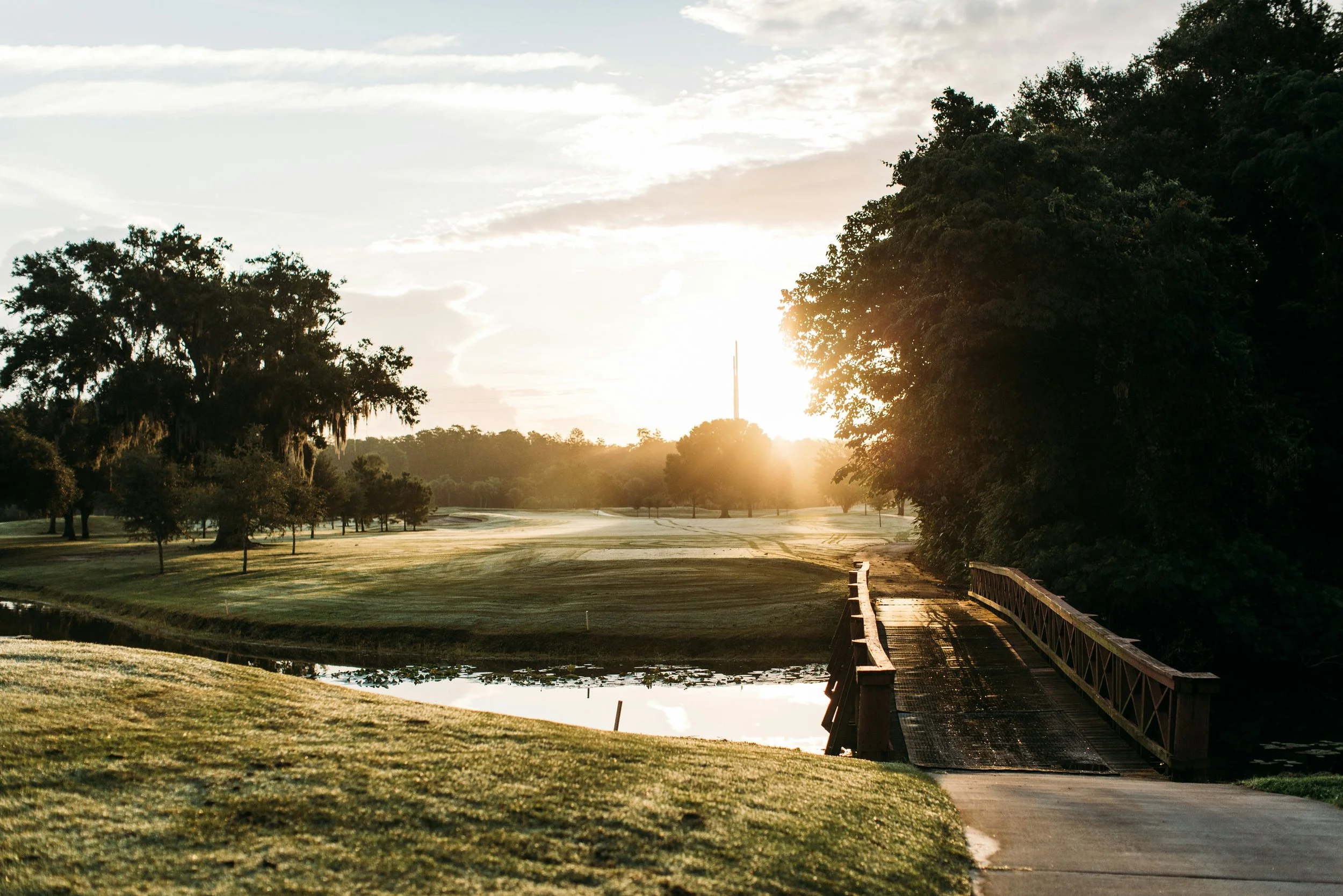 Sunset over a golf course with trees, a pond, and a wooden bridge.