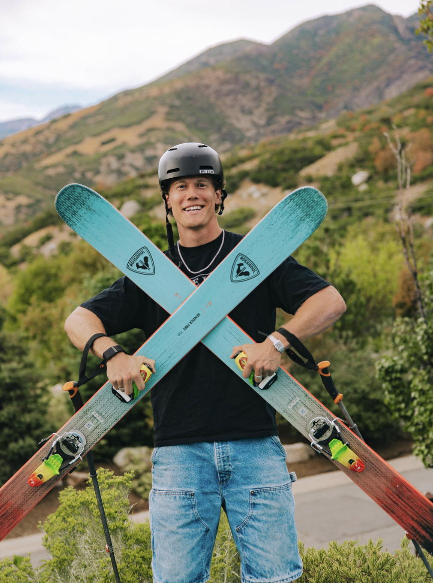 A smiling man wearing a black helmet and black T-shirt holding two crossed skis outdoors in a mountainous area with green and brown foliage.
