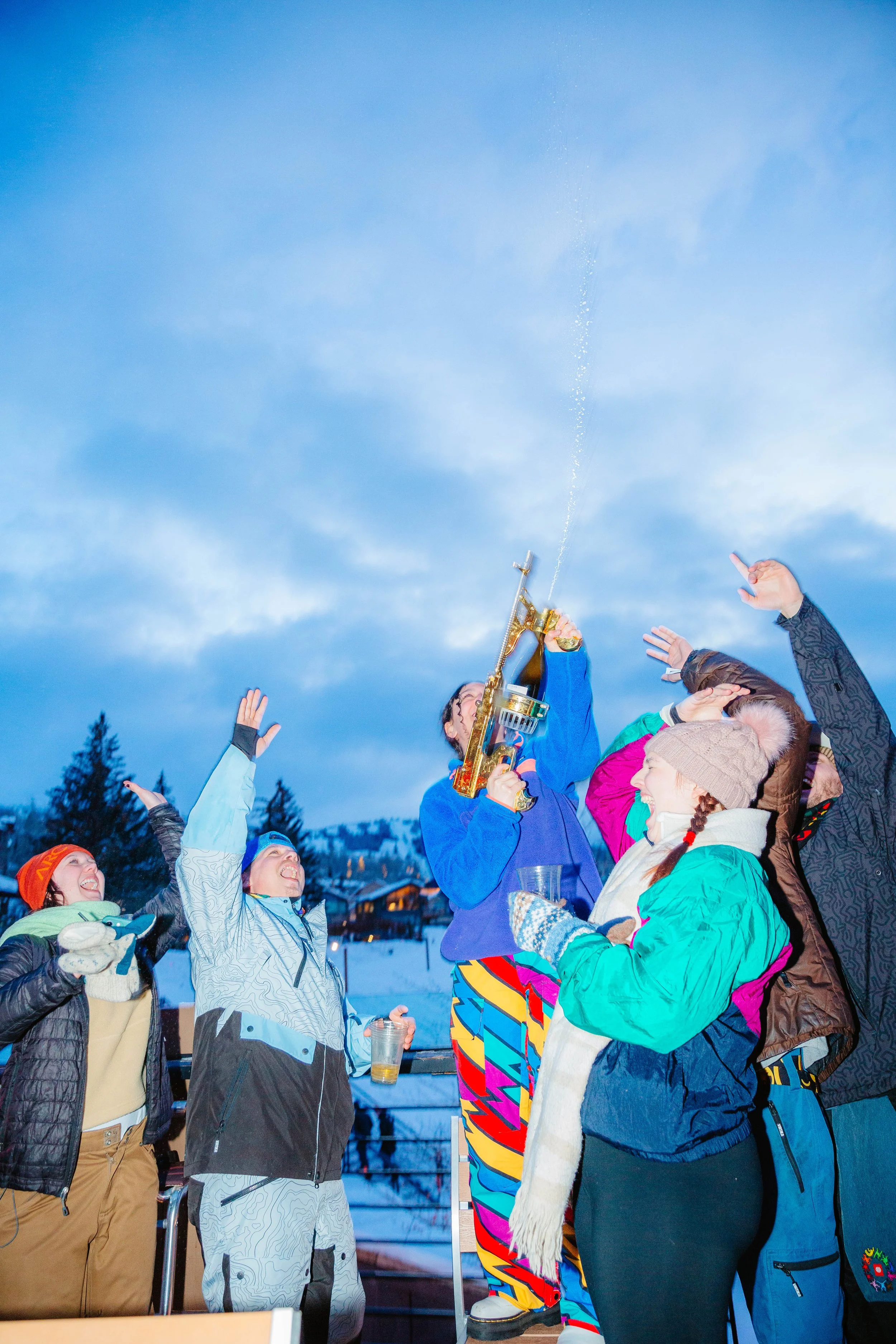 People celebrate outdoors in winter, holding glasses and raising hands as a person on the right sprays champagne from a bottle. The group is dressed in colorful winter clothing, and snow-covered trees are visible in the background.