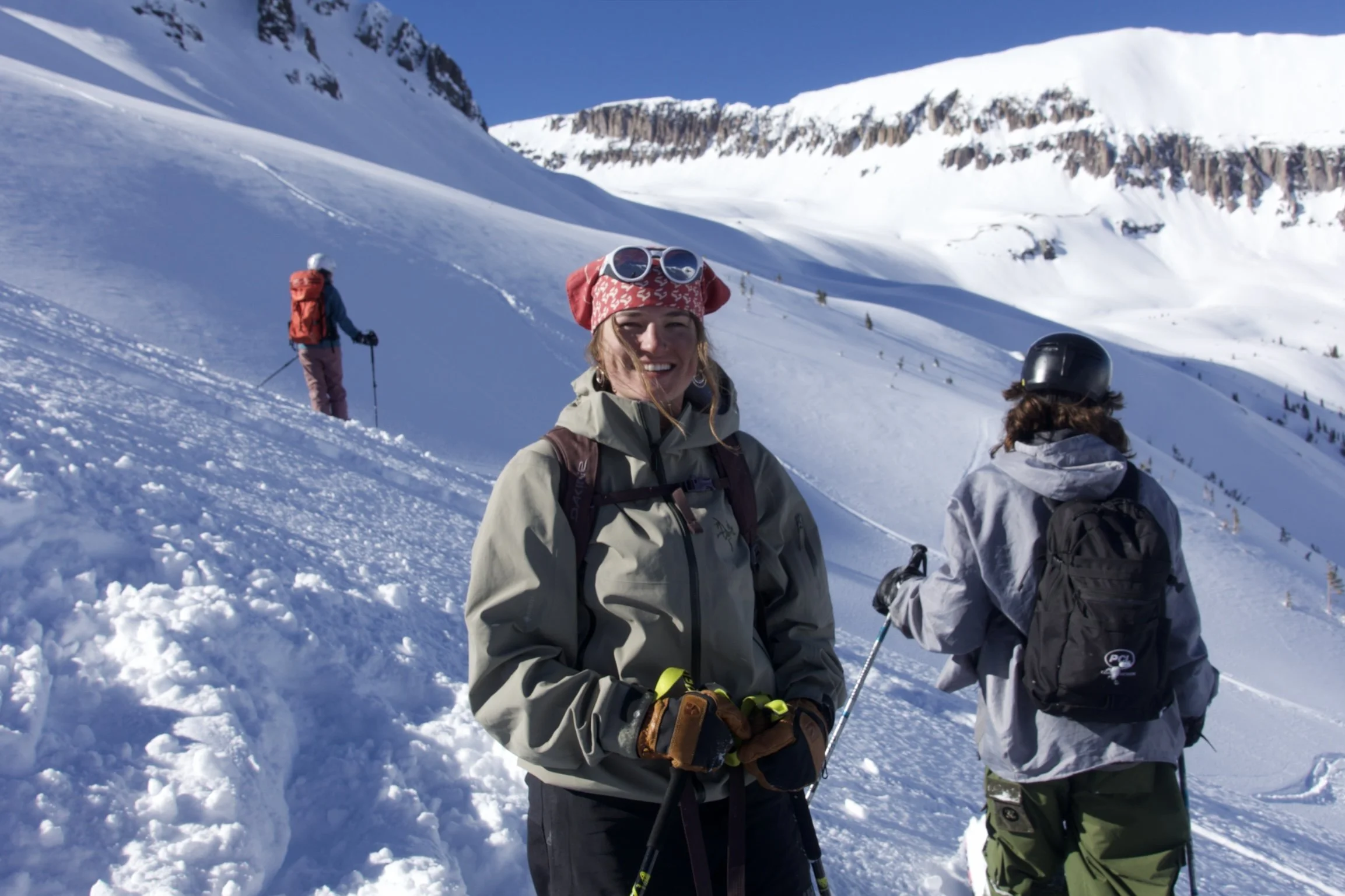 Three people are on a snowy mountain slope with a clear blue sky in the background. One woman in the foreground is smiling, wearing a red bandana with sunglasses on top, a beige jacket, and gloves. Two others are seen from behind, wearing helmets and winter gear, with one standing and the other kneeling, engaged in winter sports activities.