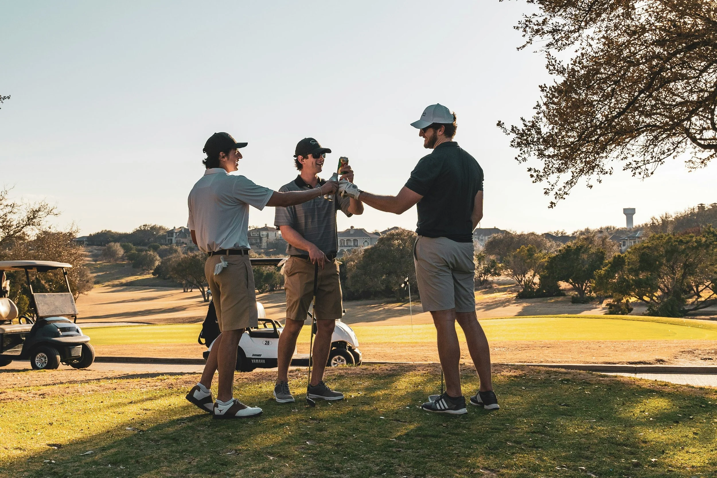 Three men are on a golf course, sharing a drink and talking, with golf carts and trees in the background during the daytime.