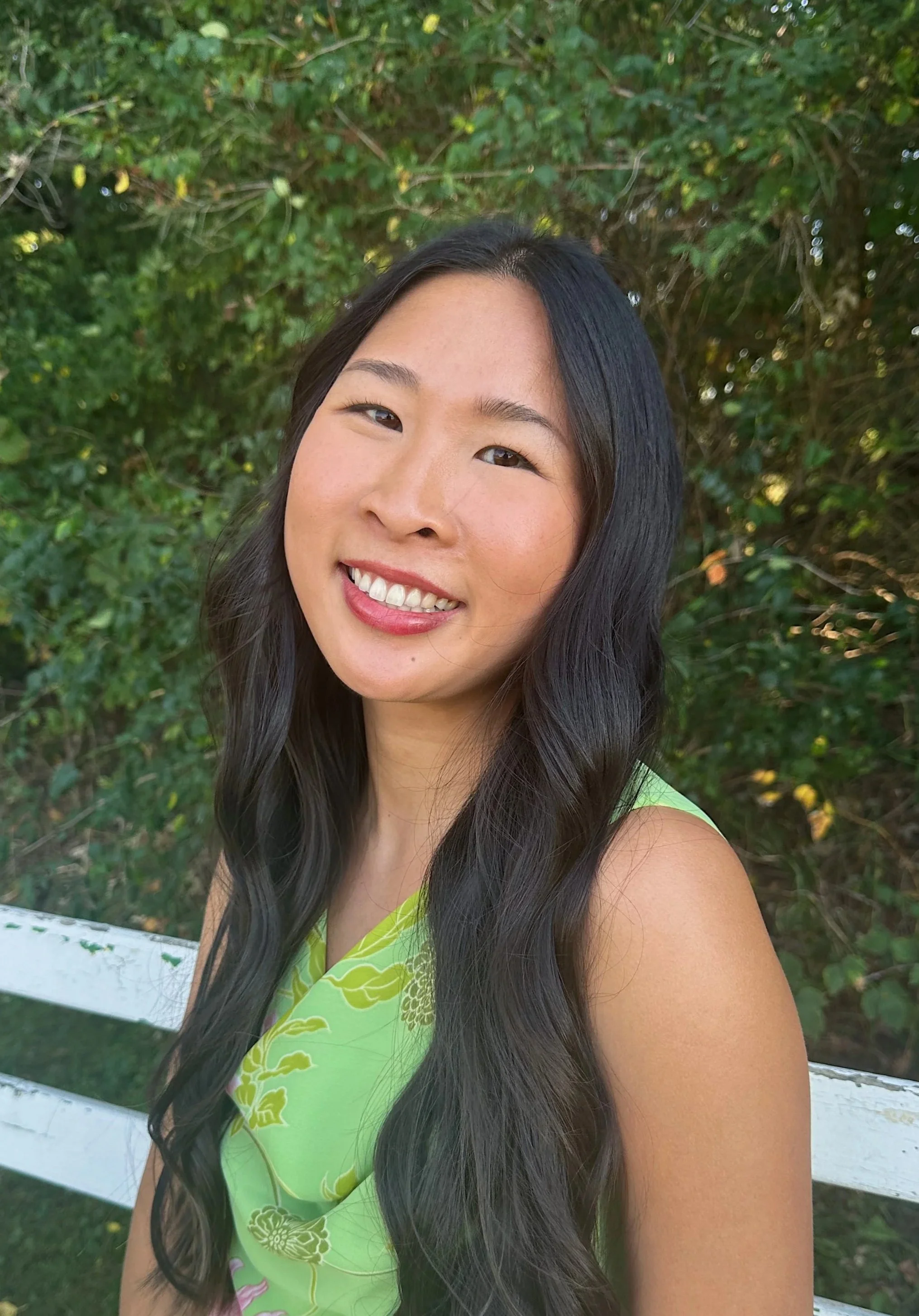 A young woman with long black wavy hair smiling outdoors with green foliage and a white fence in the background.