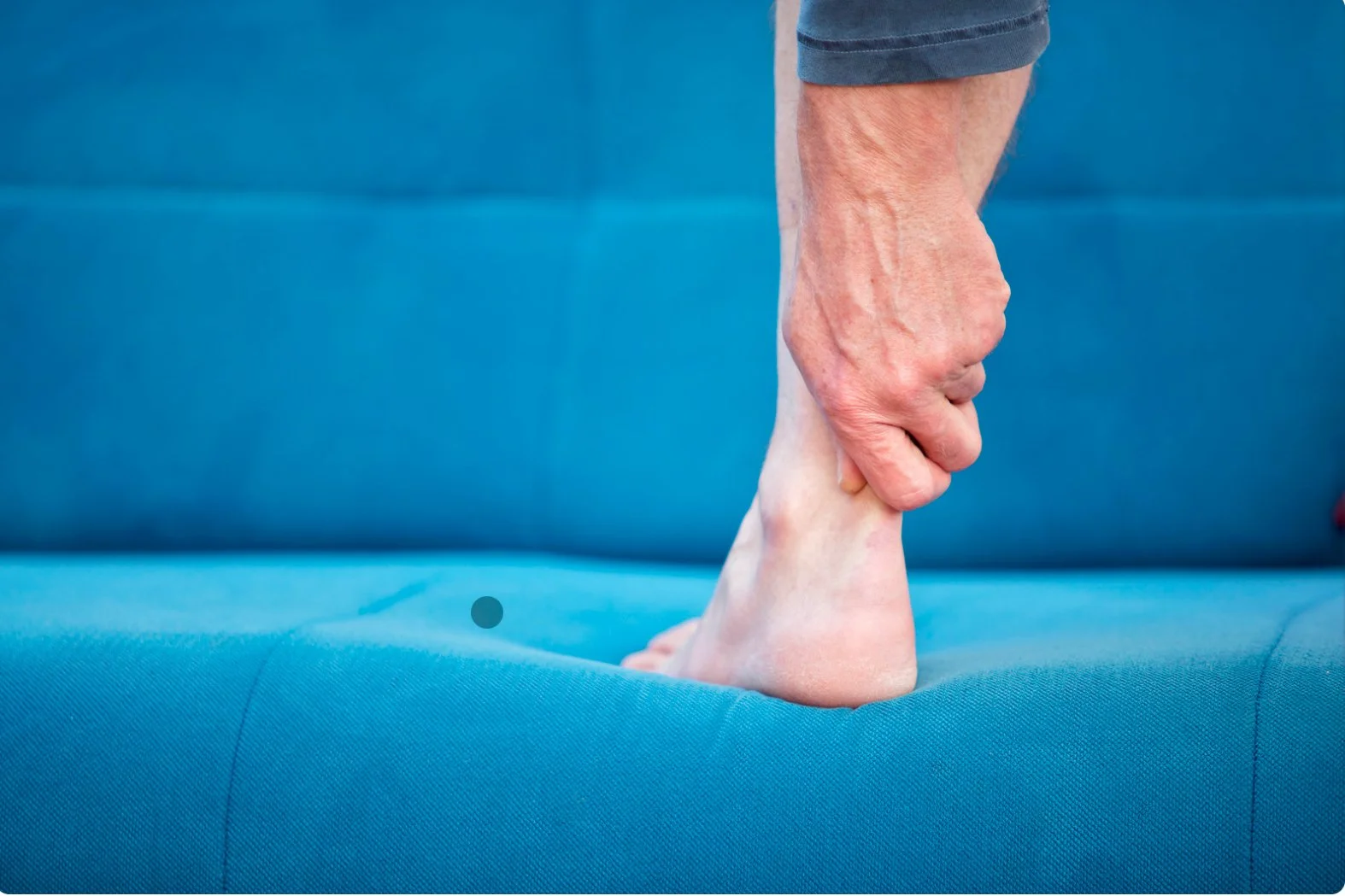 Close-up of a person's hand gripping their ankle while standing on a blue couch or bed with achilles tendonitis.