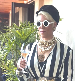 Woman wearing striped blazer, sunglasses, and multiple necklaces, holding a glass of champagne, standing next to green plants.