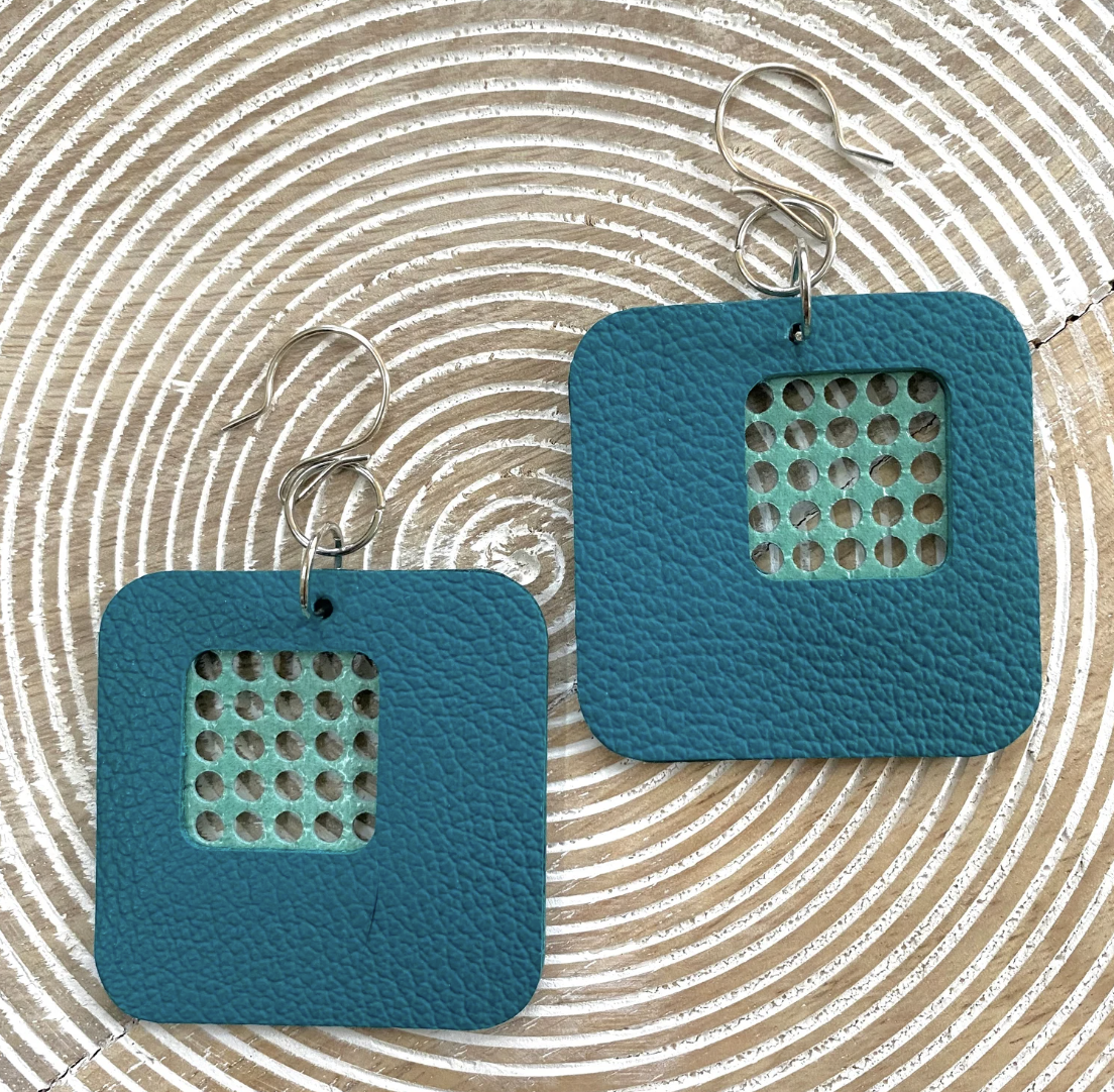 Pair of bright blue square earrings with perforated grid patterns in the center, resting on a textured circular wooden surface.