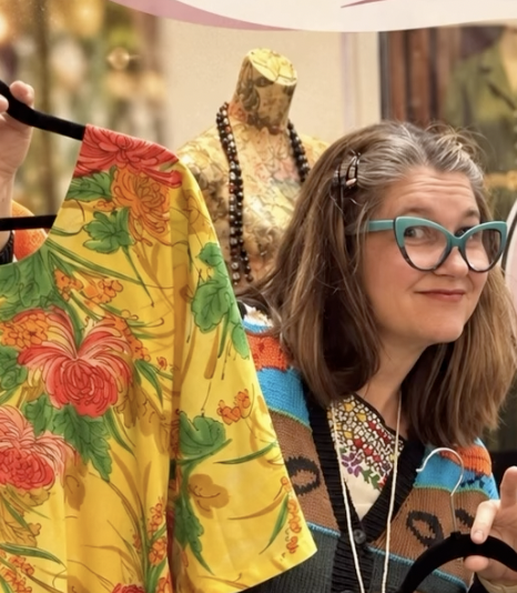 Woman with glasses in a colorful sweater, standing near a mannequin dressed in a yellow floral garment, inside a store.