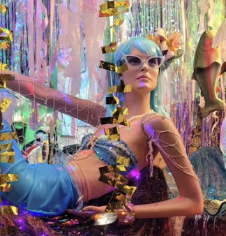 Person dressed in colorful, futuristic fashion with blue hair, wearing large white sunglasses, surrounded by shiny decorations and colorful streamers.
