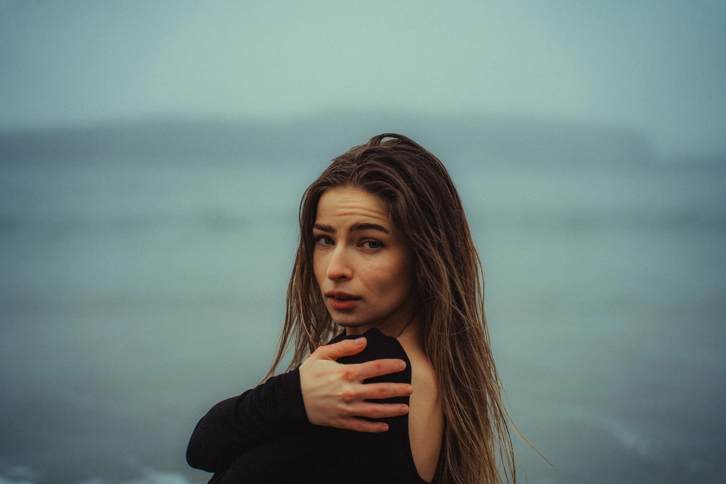 A young woman with long brown hair stands outdoors near a body of water, wearing a black top, with her hand on her shoulder, looking over her shoulder towards the camera.