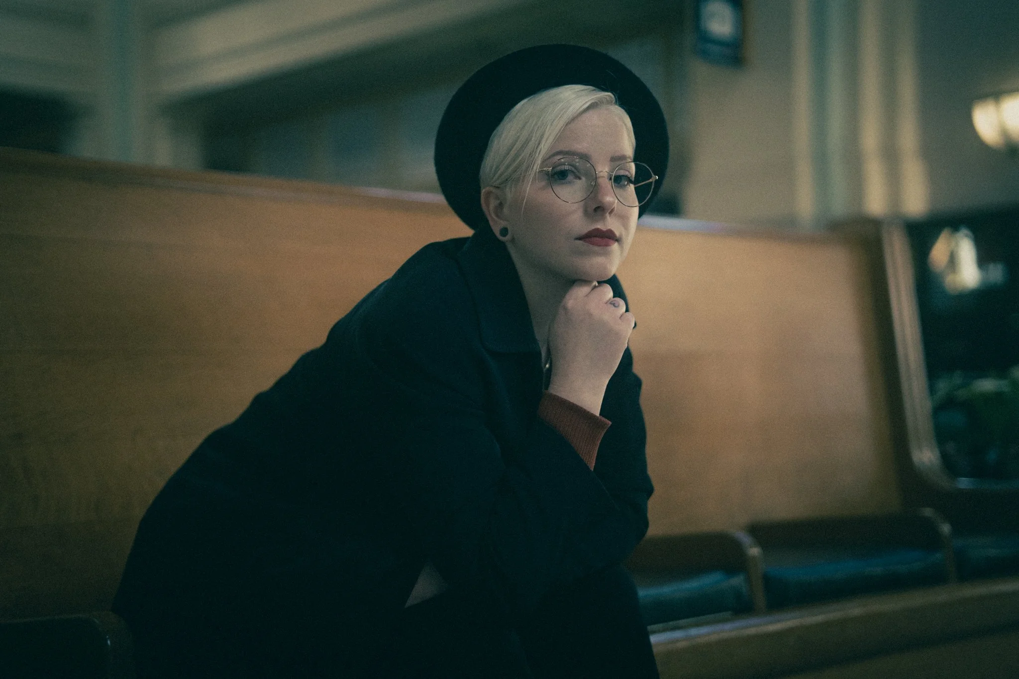 A woman with short blonde hair, wearing round glasses, a black hat, and a black jacket, sitting on a wooden bench in what appears to be a vintage-style train station or waiting area.