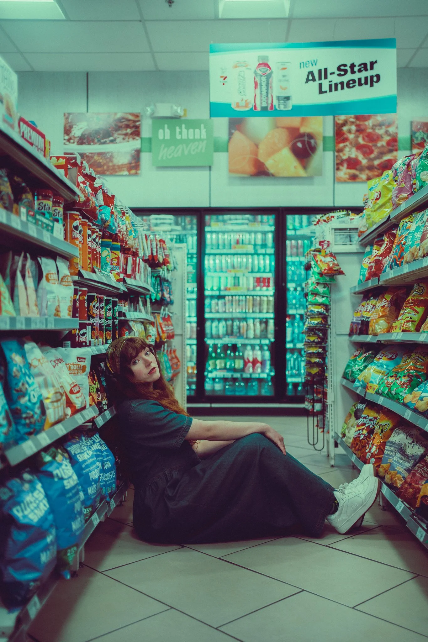 A woman with long hair and bangs is sitting on the floor in a grocery store aisle between shelves of snacks, looking up at the camera with a relaxed expression.