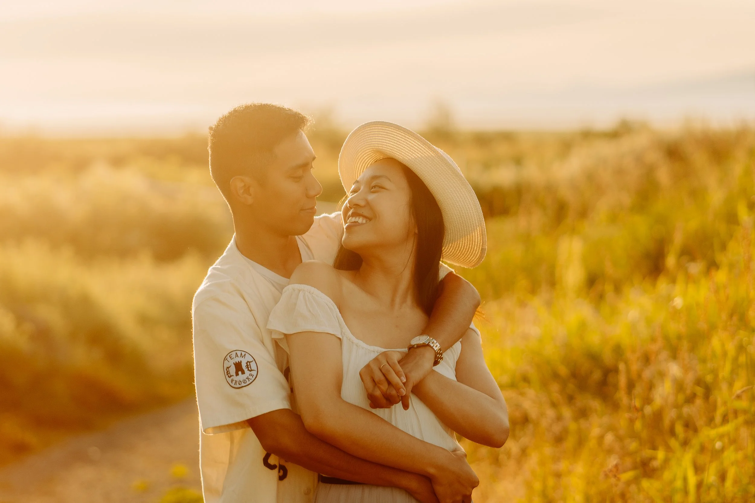 A couple embraces in a field during sunset, with the man wearing a light shirt and the woman wearing a wide-brimmed hat and a white dress, smiling at each other.
