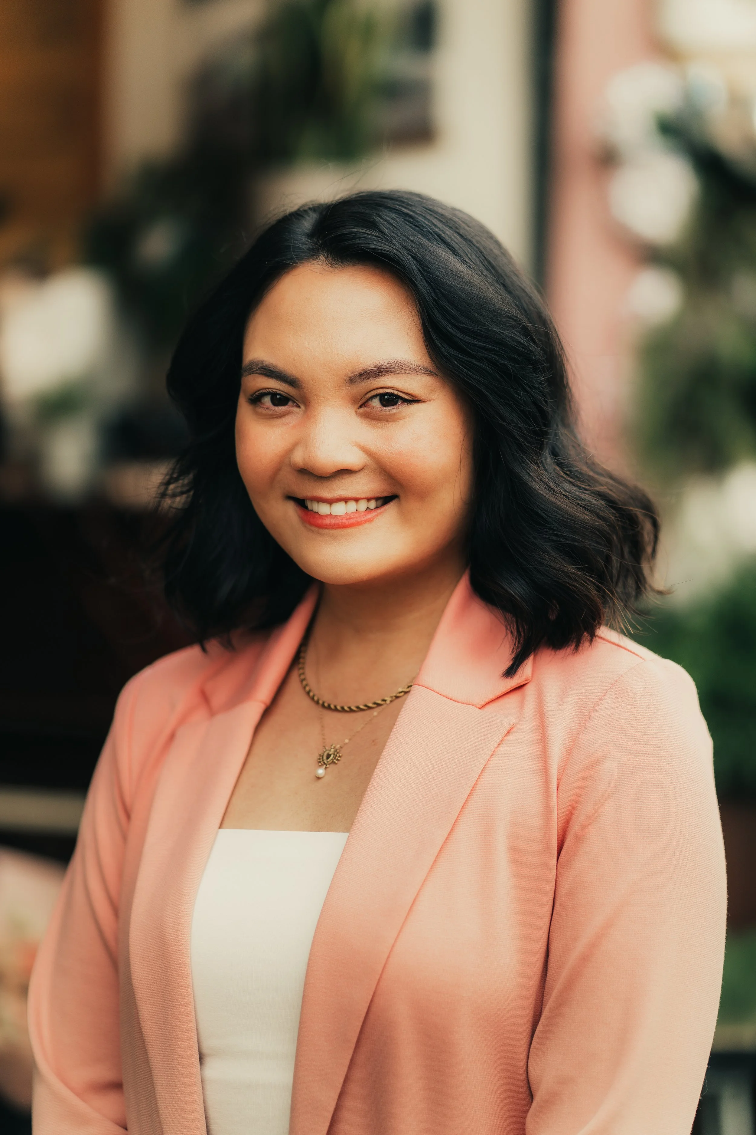 A woman with shoulder-length black hair, smiling, wearing a light pink blazer and white top, standing outdoors with a blurred background.