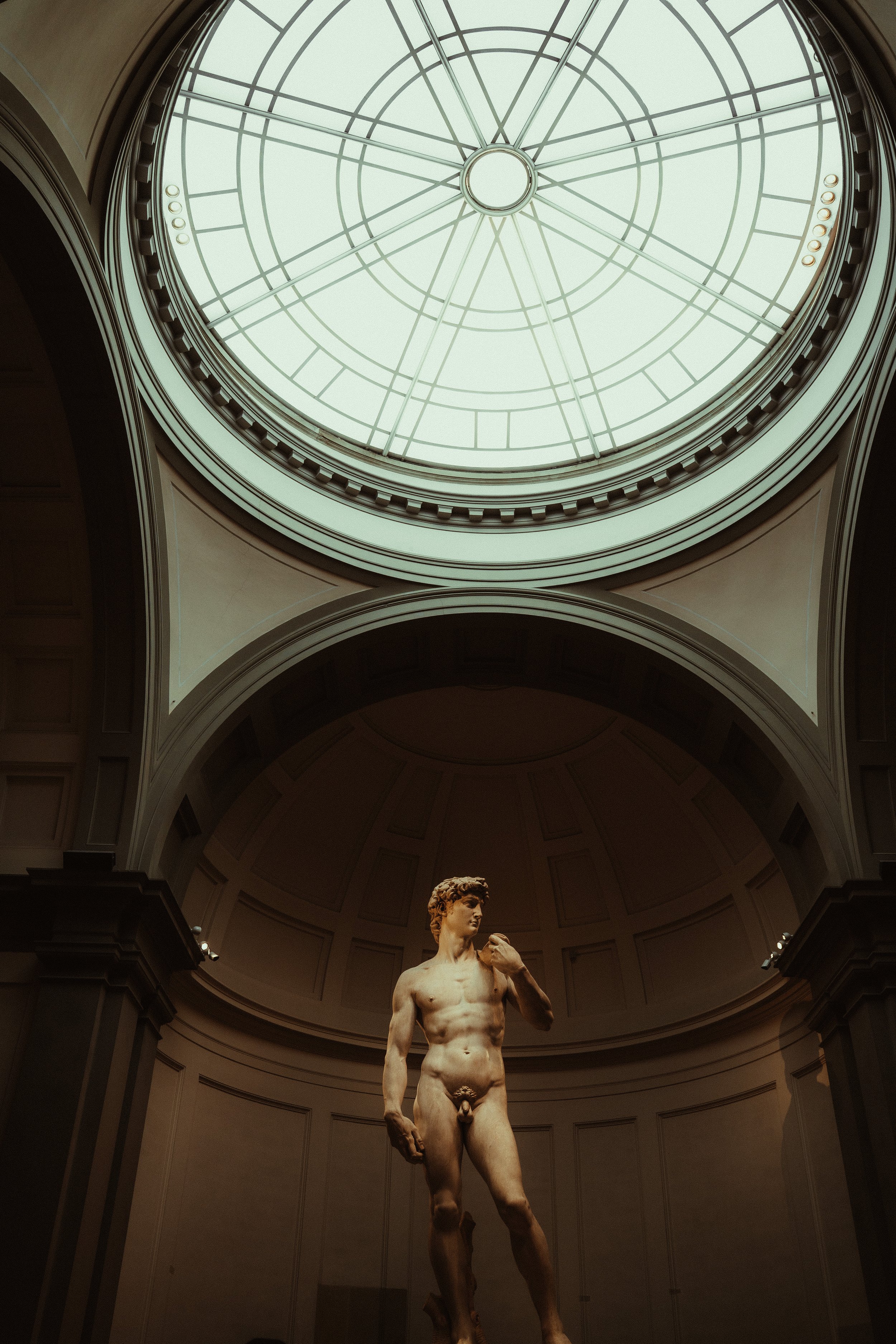 Classical sculpture of a nude male standing with a removed left hand, in a grand museum hall with a large clock face ceiling.